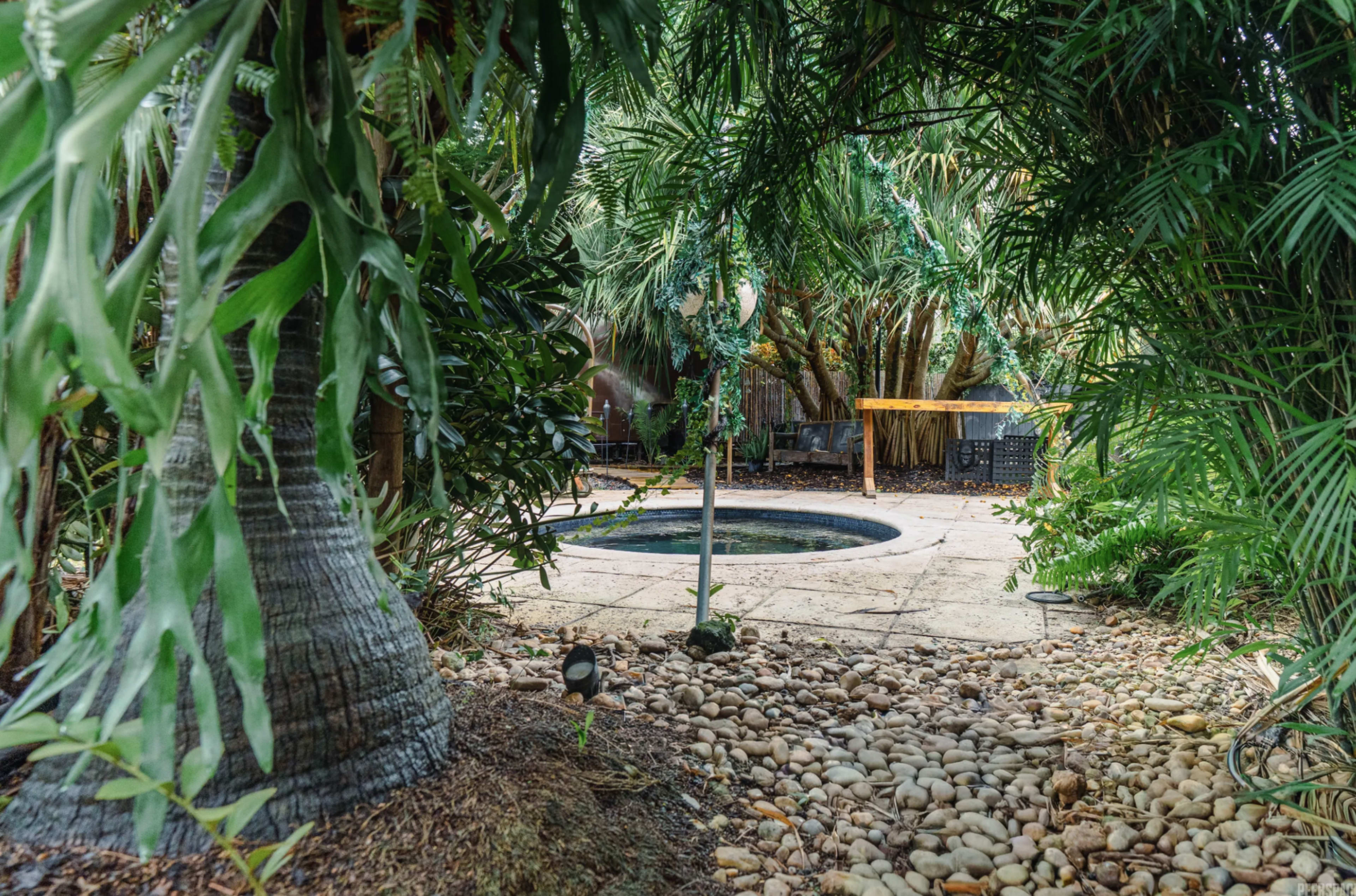 A circular hot tub is partially visible through tropical vegetation and a stone pathway in a secluded outdoor setting.