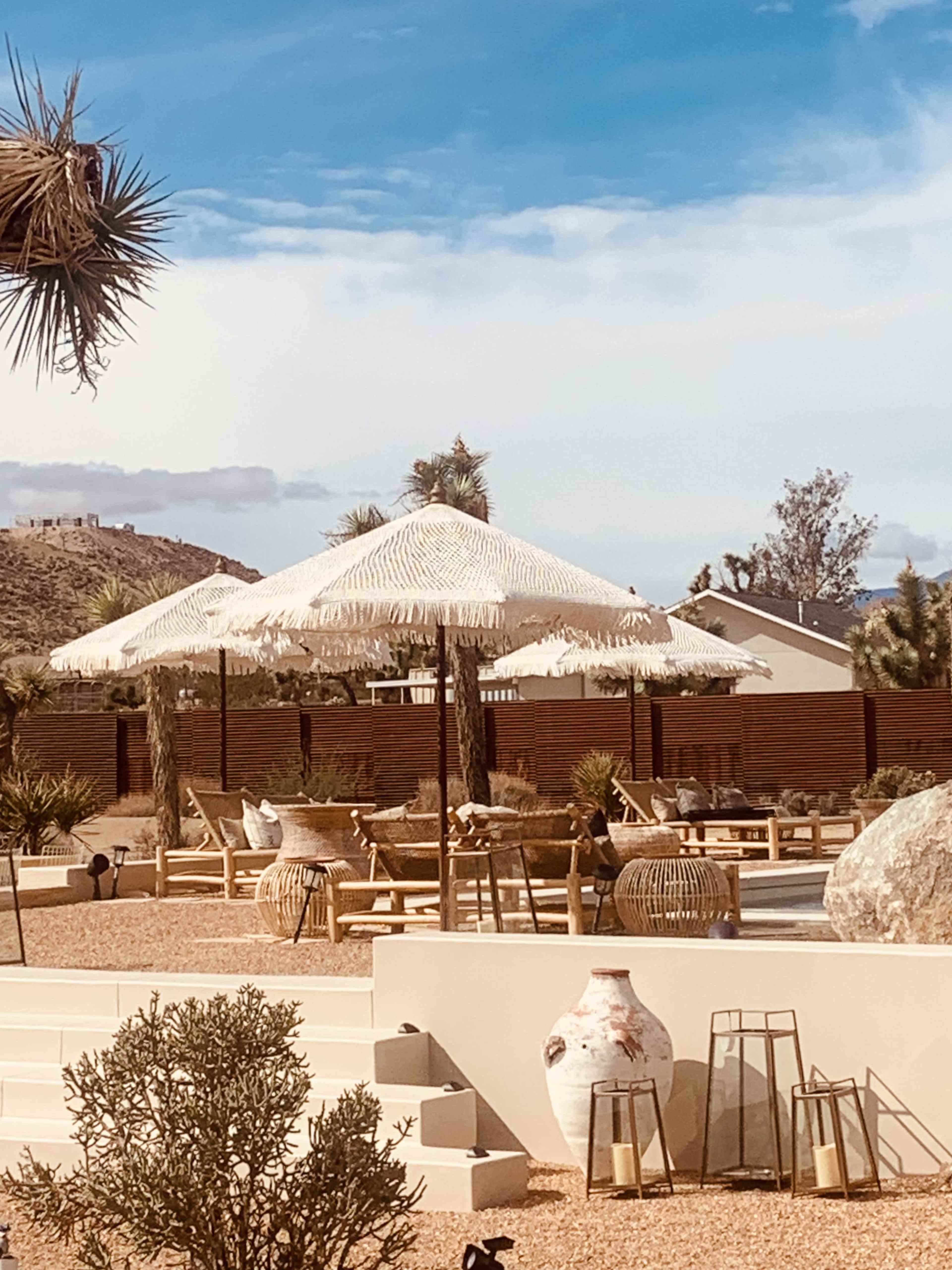 A desert landscape featuring a seating area with straw umbrellas, surrounded by cacti and decorative pottery.