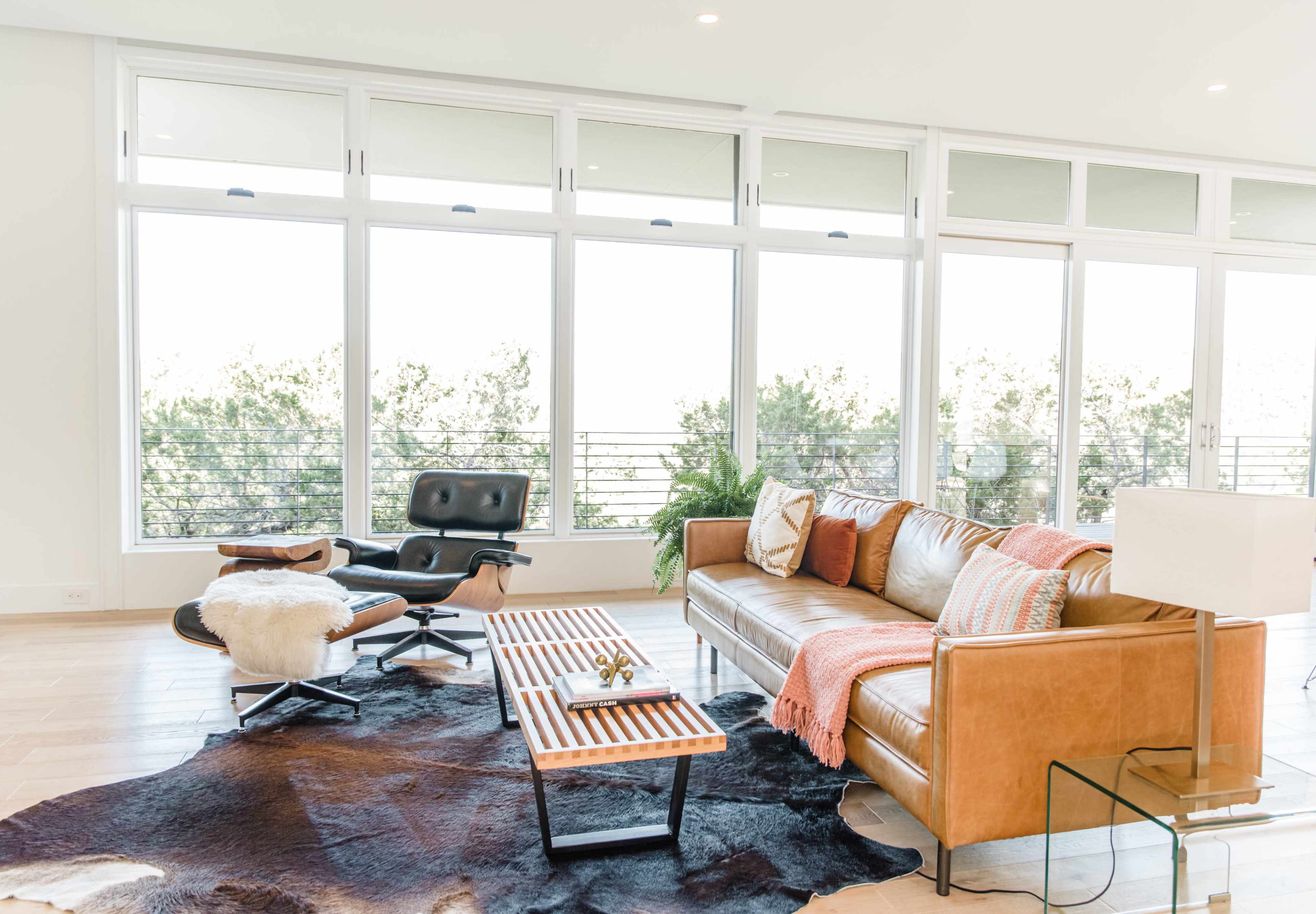 A modern living room features a leather sofa, an Eames chair, a wooden coffee table, and a cowhide rug, with large windows allowing natural light and a view of greenery outside.