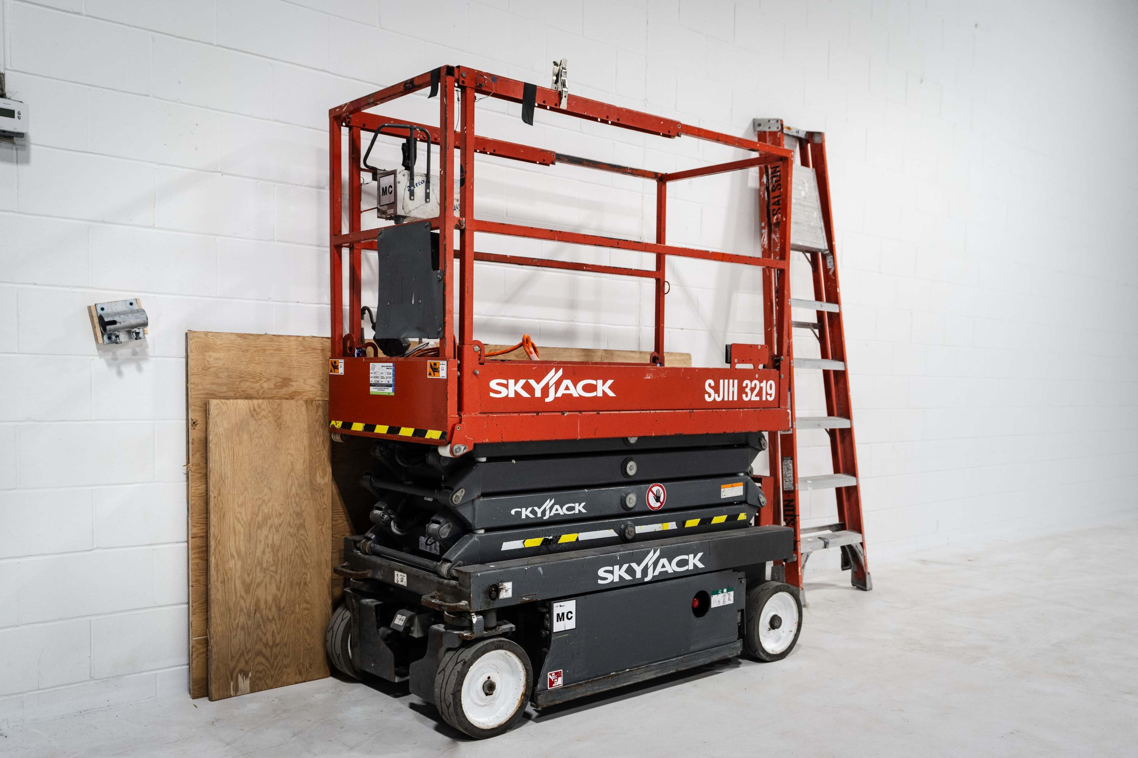 A red Skyjack scissor lift is parked next to a wooden panel and a ladder against a white wall.