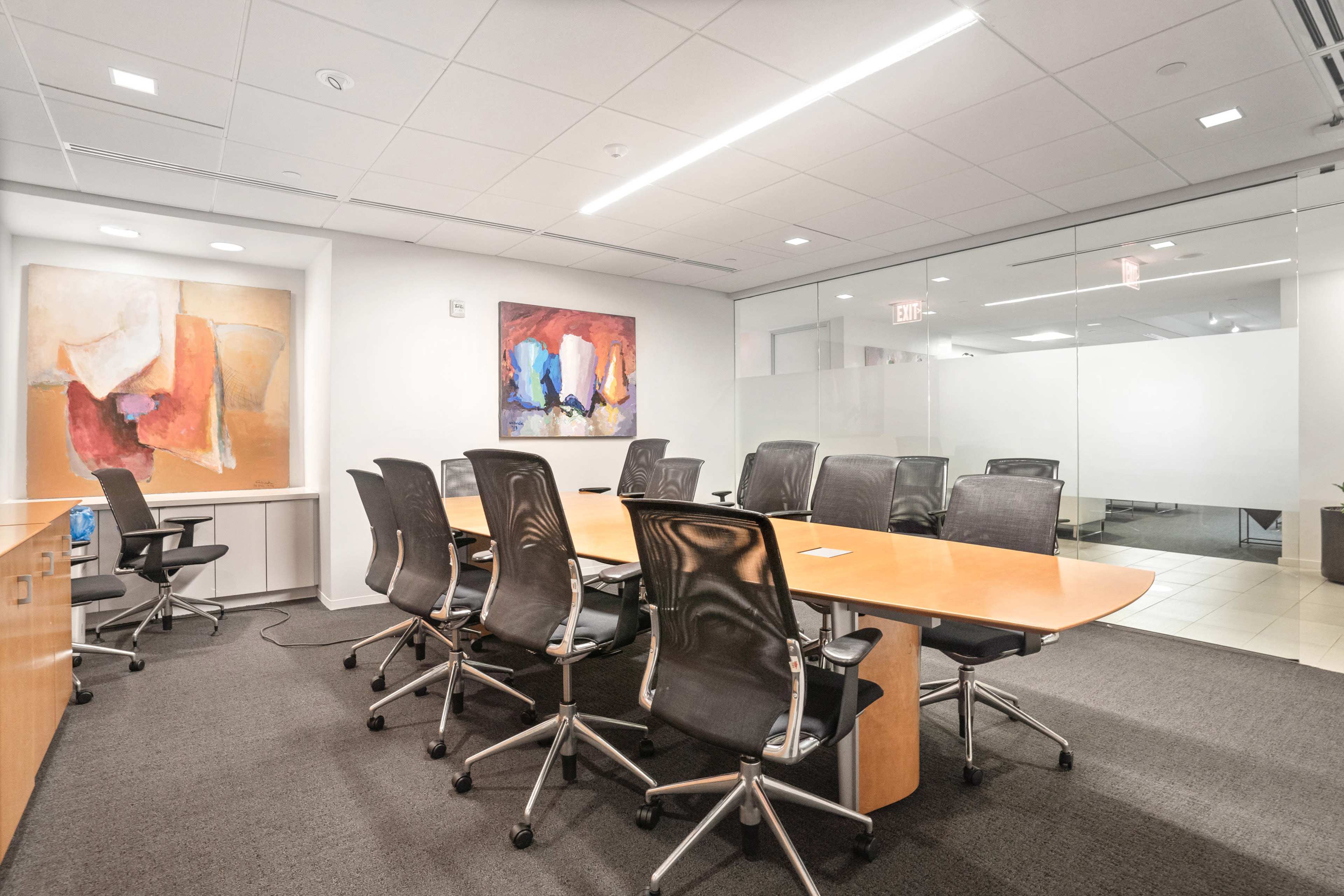 A modern conference room features a long wooden table surrounded by black office chairs, with two colorful abstract paintings on the walls.