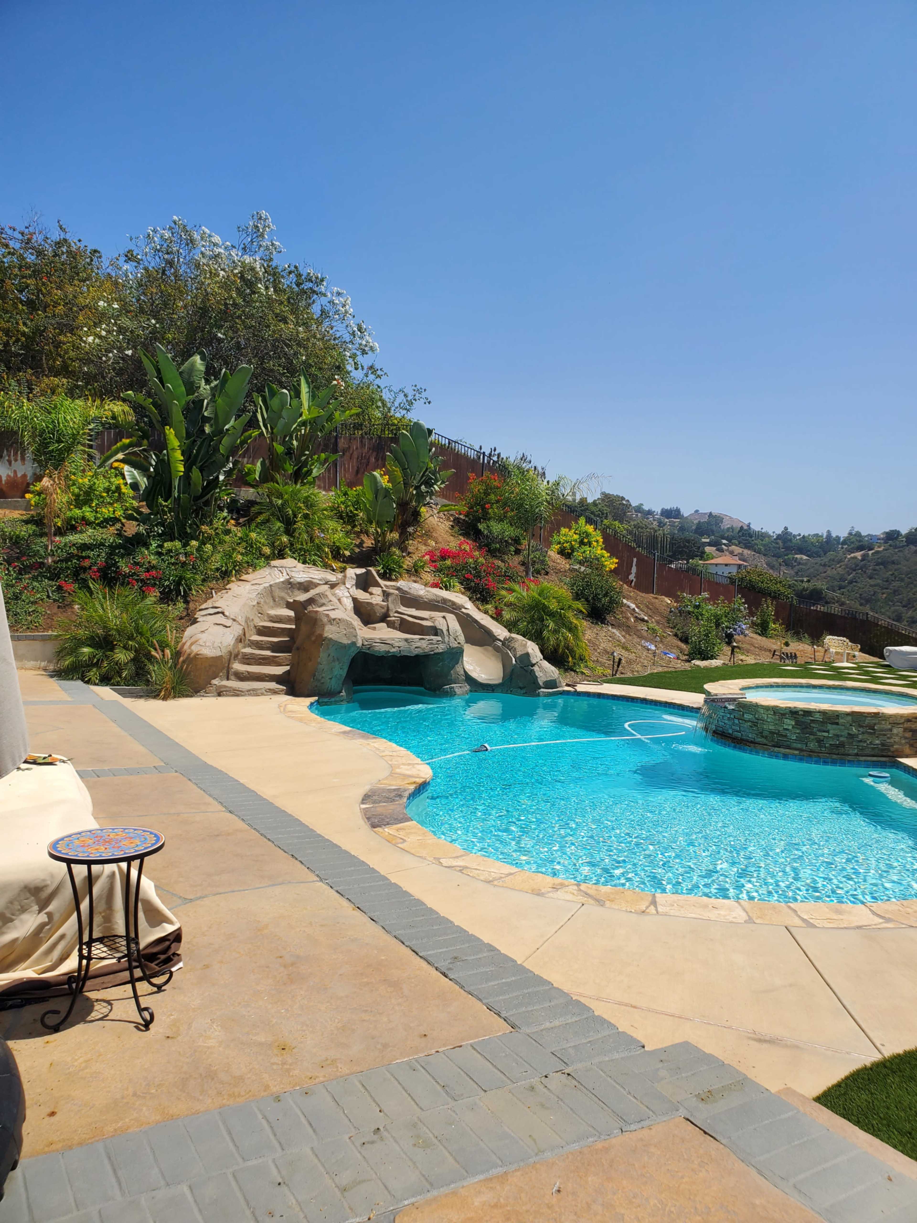 A landscaped backyard with a swimming pool featuring a rock waterfall and a hot tub, surrounded by greenery and colorful flowers.