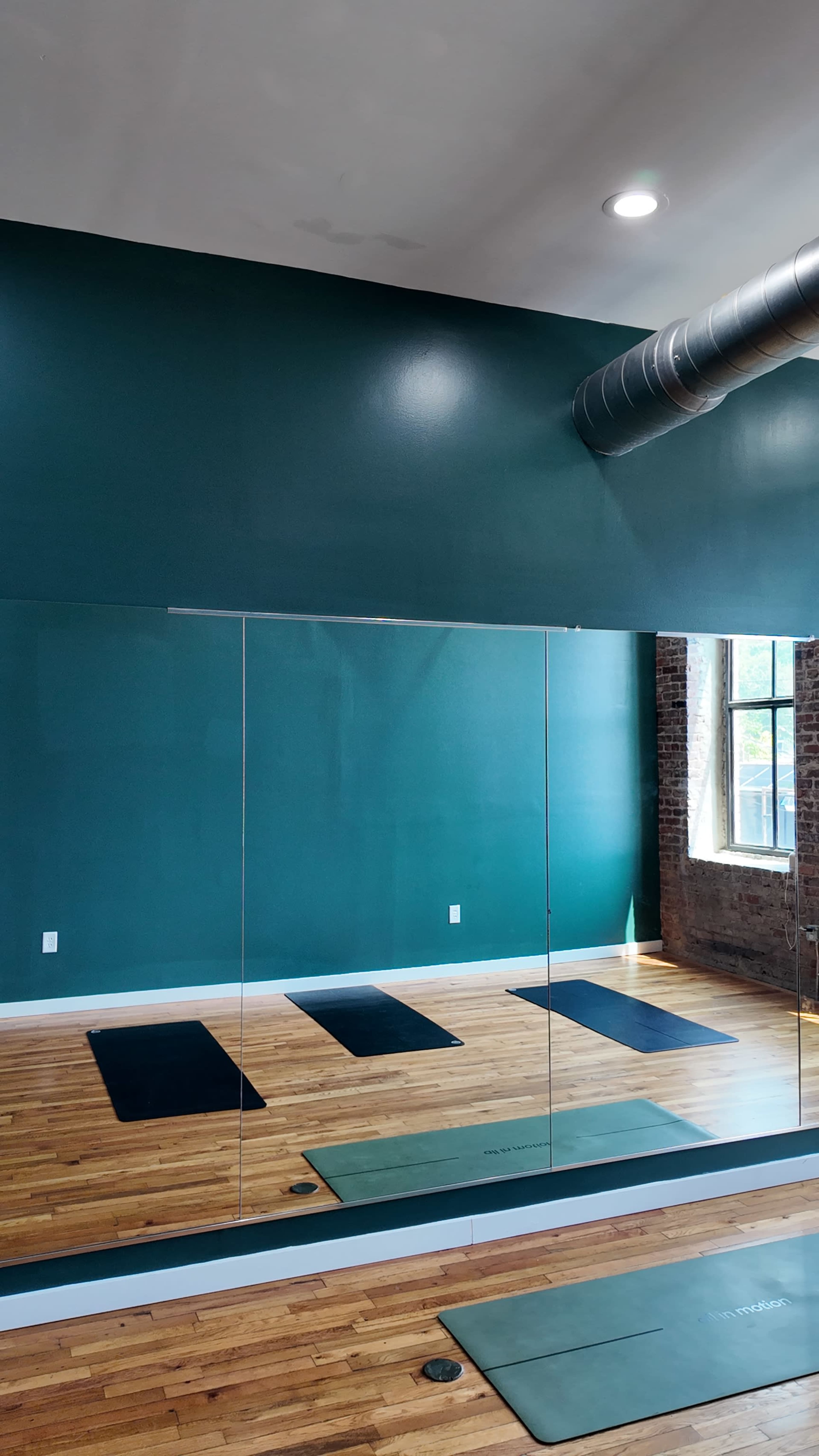 The image shows a yoga studio with wooden flooring, four yoga mats placed on the floor, and a large mirror reflecting a teal wall and a window with exposed brick.