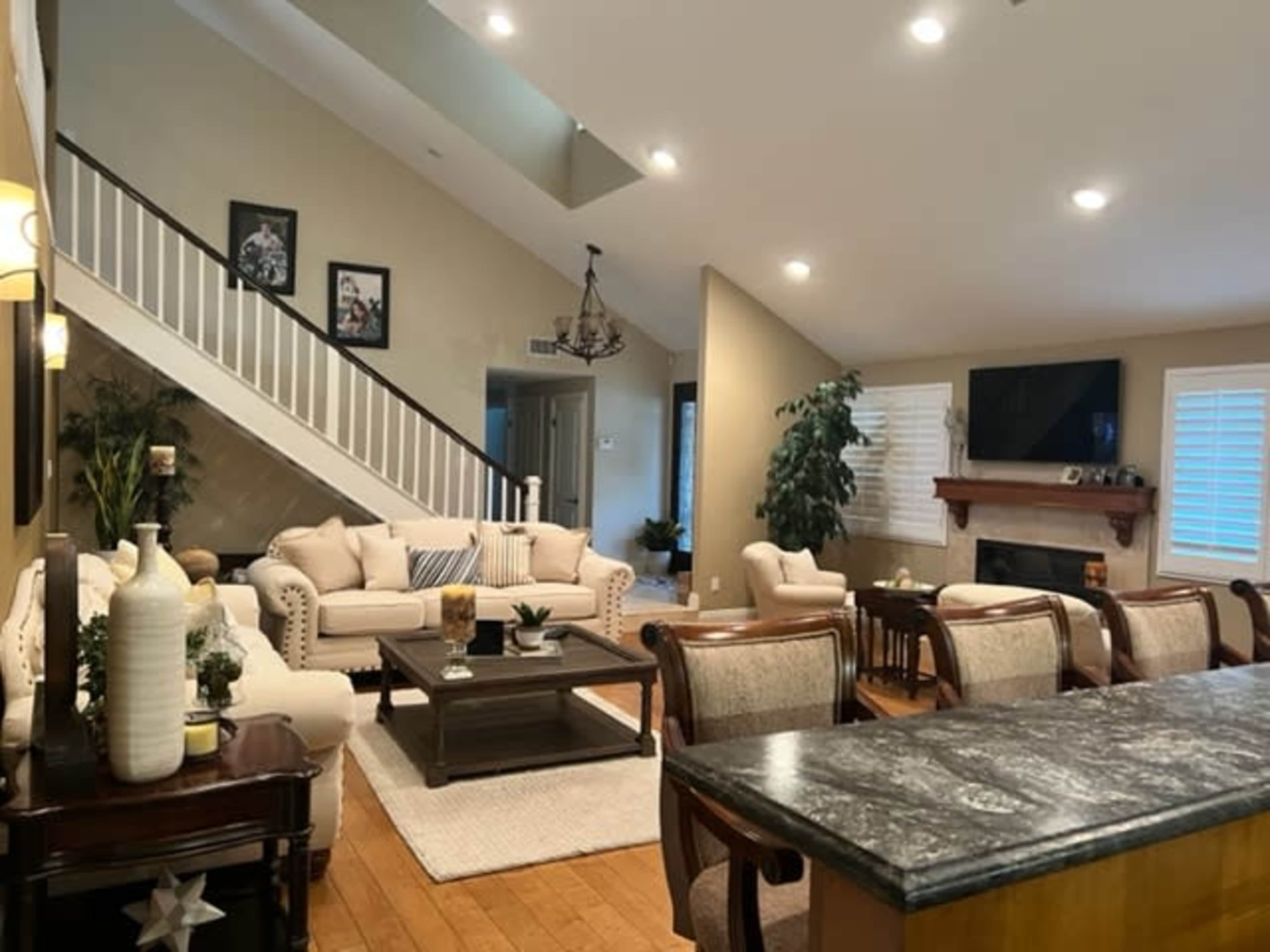 A spacious living area with a staircase, a light-colored sofa set, a wooden dining table, and a fireplace.