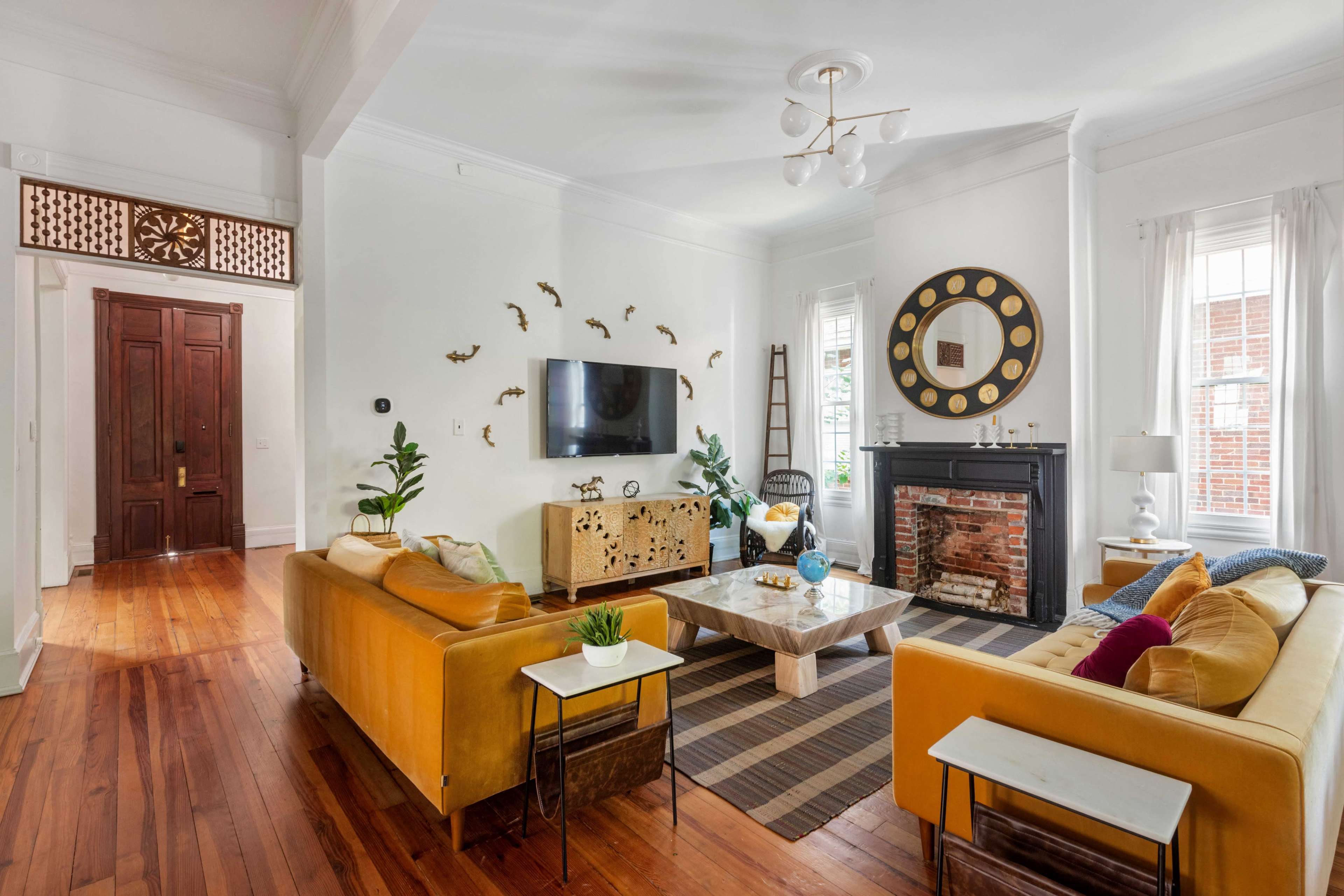 The living room features a combination of yellow sofas, a coffee table, a television mounted on the wall, and a fireplace, with large windows allowing natural light to illuminate the space.
