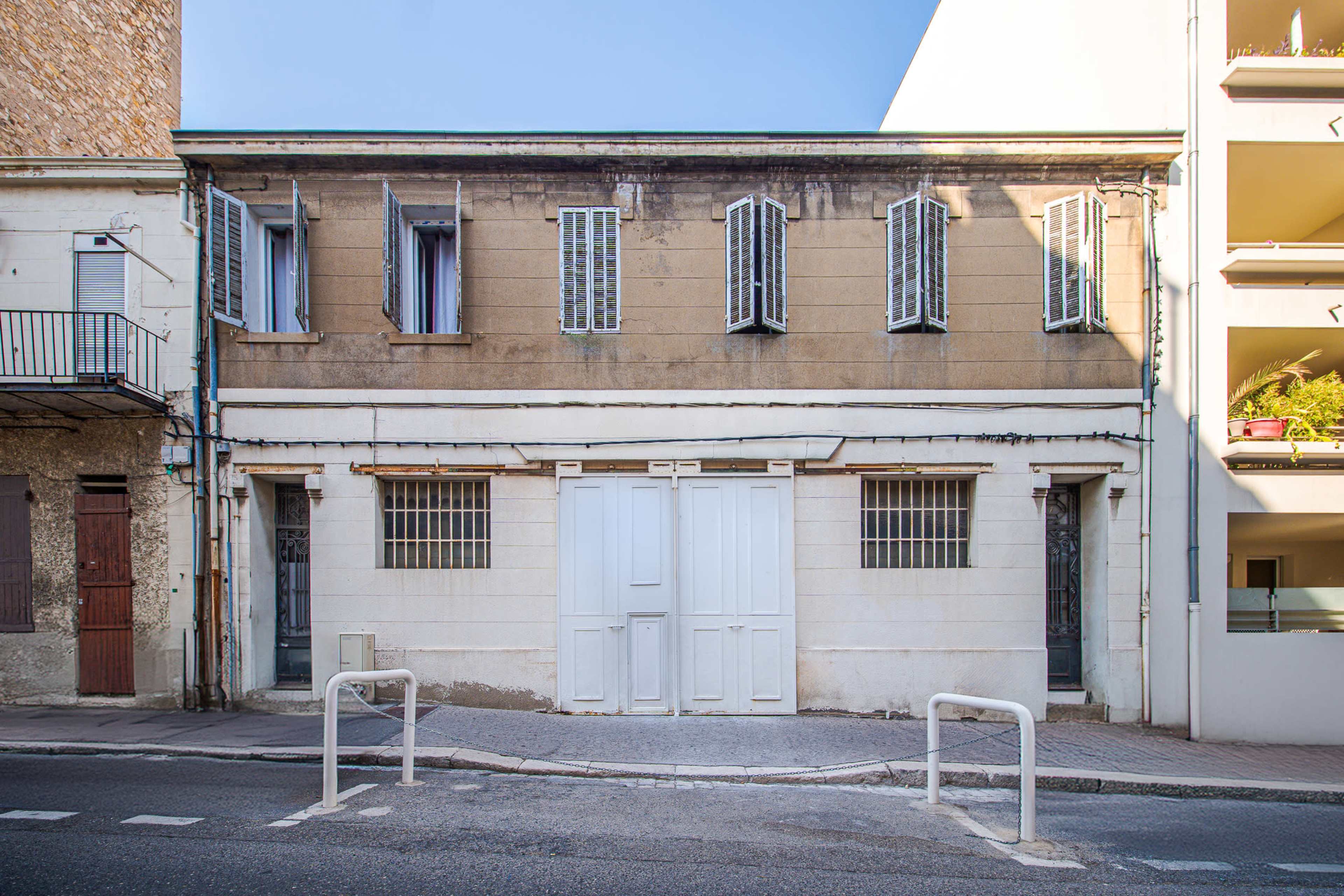 The image shows a two-story building with a stone facade, white doors, and multiple shuttered windows, situated on a street next to a modern apartment complex.