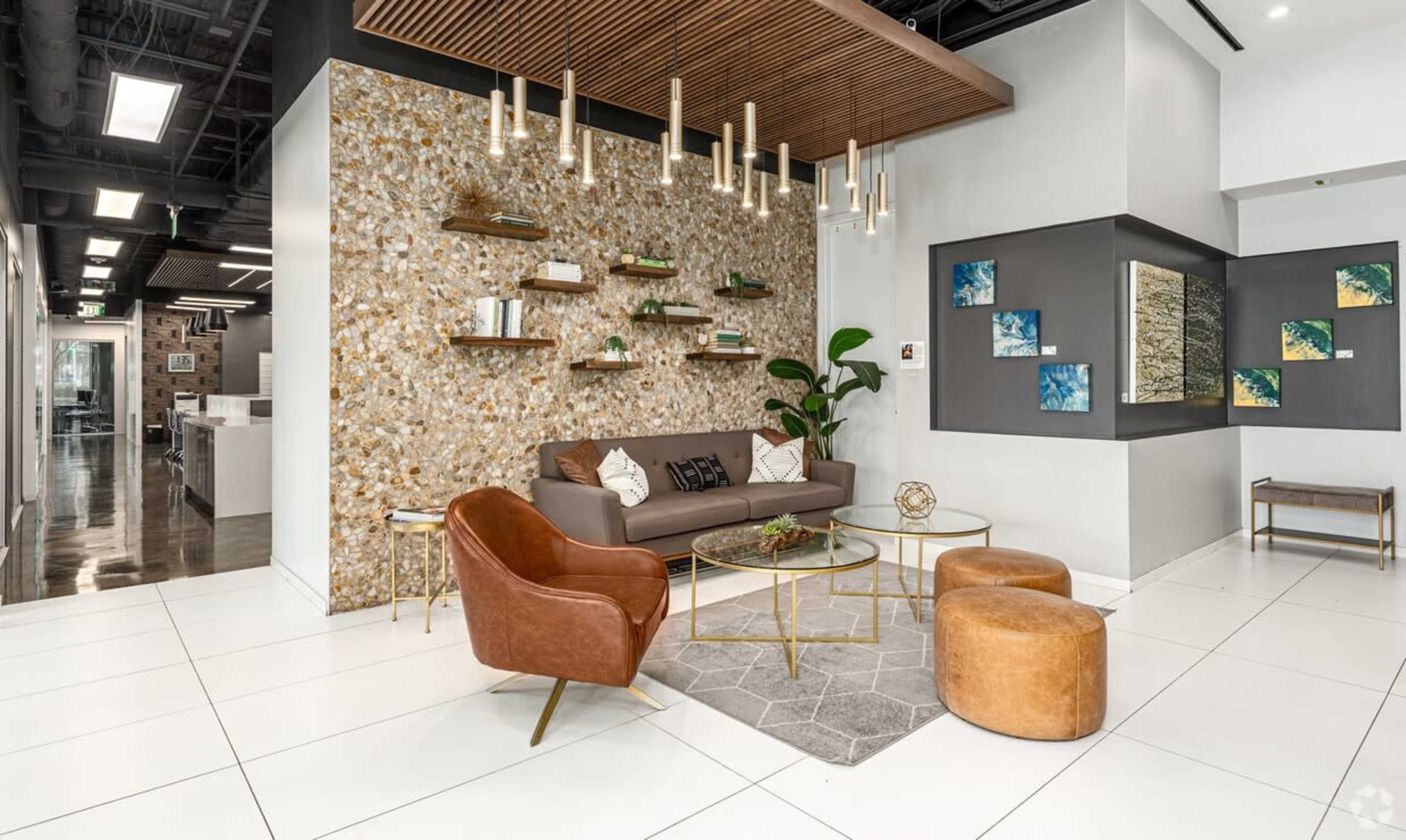 The image depicts a modern lounge area with a brown sofa, decorative shelving on a textured wall, and gold accents, surrounded by circular wooden tables and greenery.