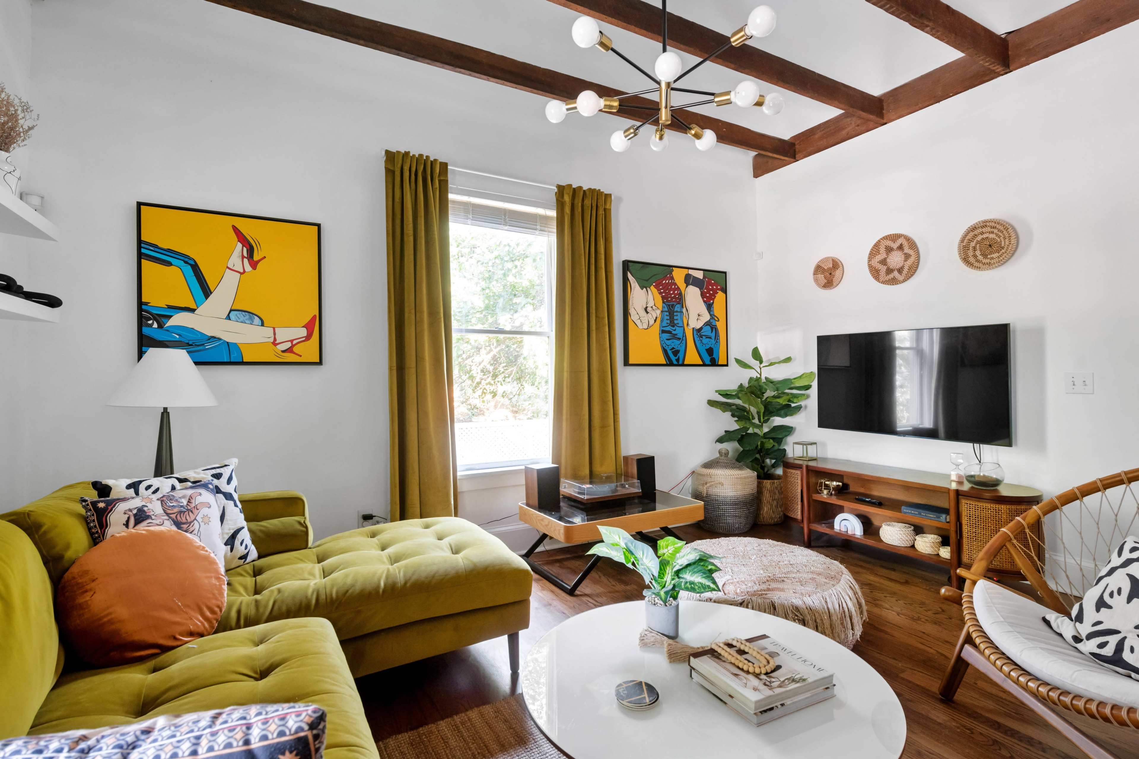 The living room features a bright yellow sofa, two framed artworks, large window curtains, and a variety of furniture including a round coffee table and a TV stand.