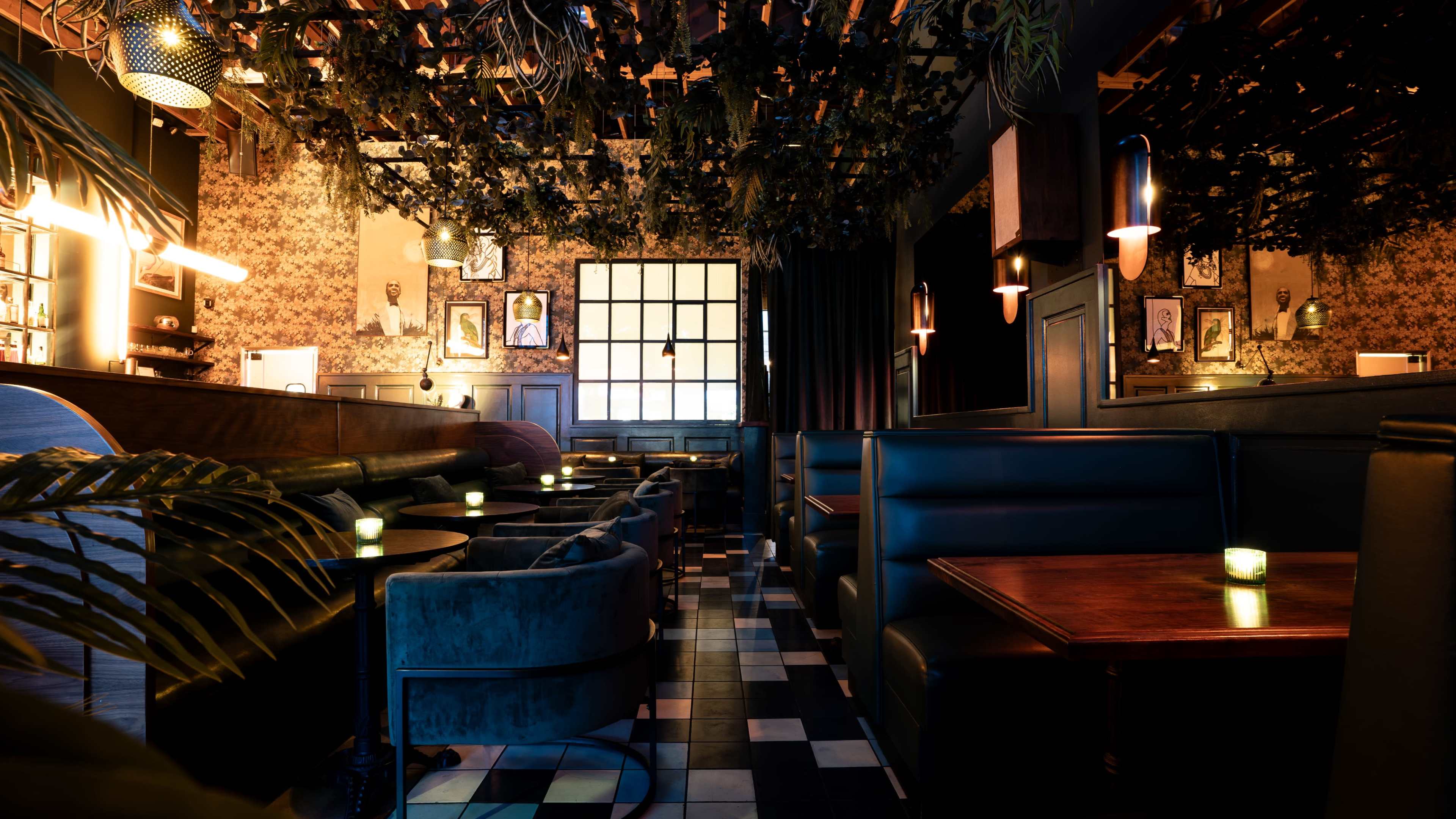 The image depicts a dimly lit restaurant interior featuring dark booths, wooden tables, and greenery hanging from the ceiling.