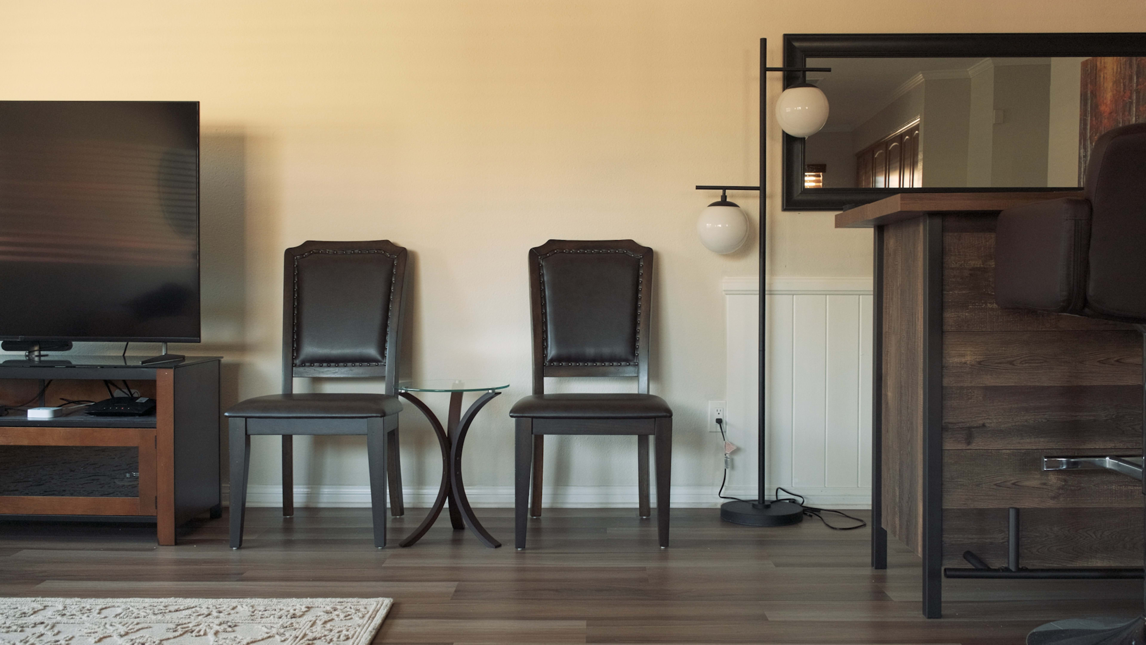 A living room setup featuring two dark leather chairs, a small glass side table, a floor lamp with two globes, a wooden bar counter, and a television unit against a light-colored wall.