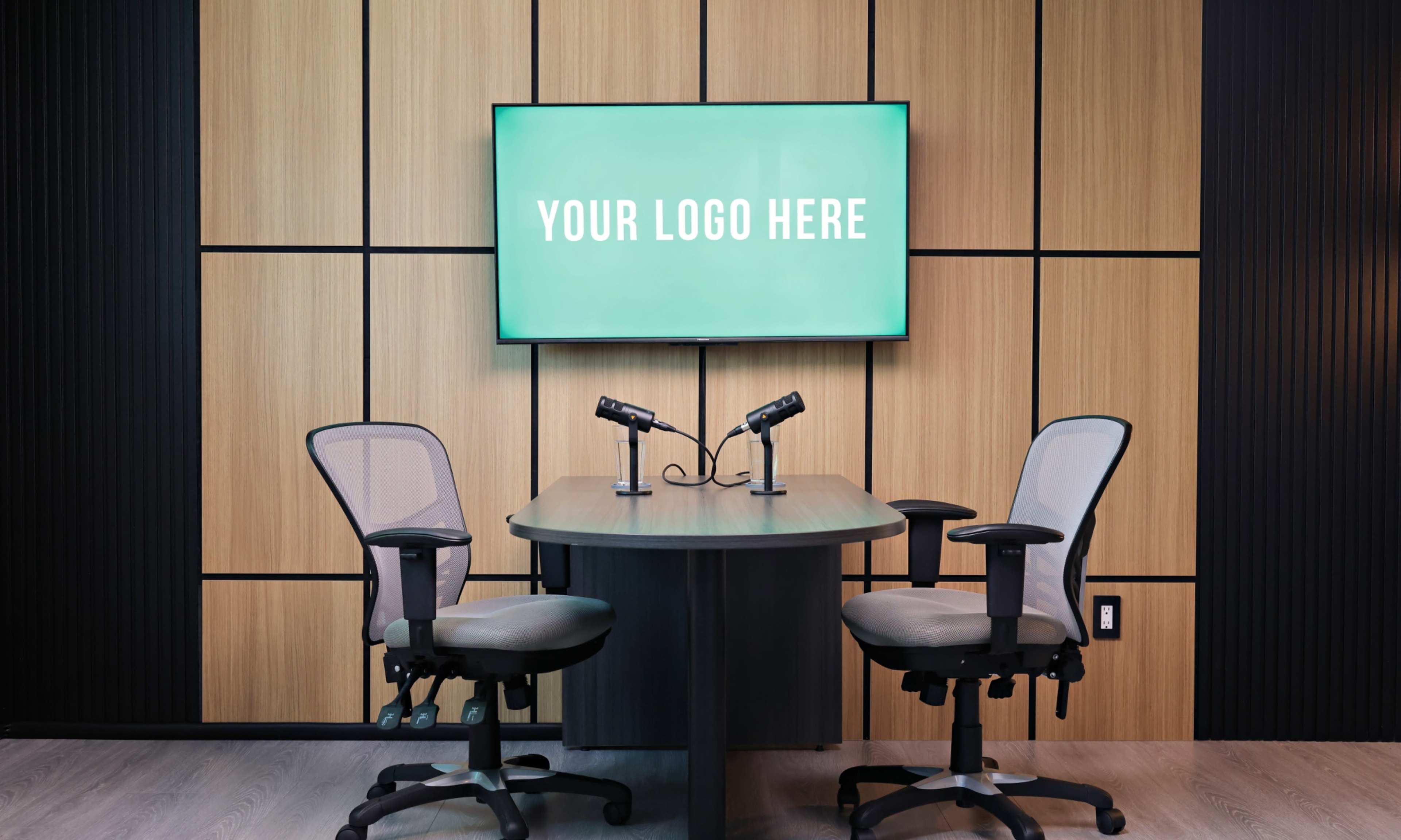The image shows a modern conference room with two grey office chairs positioned at a round table, and a digital screen on the wall displaying placeholder text for a logo.
