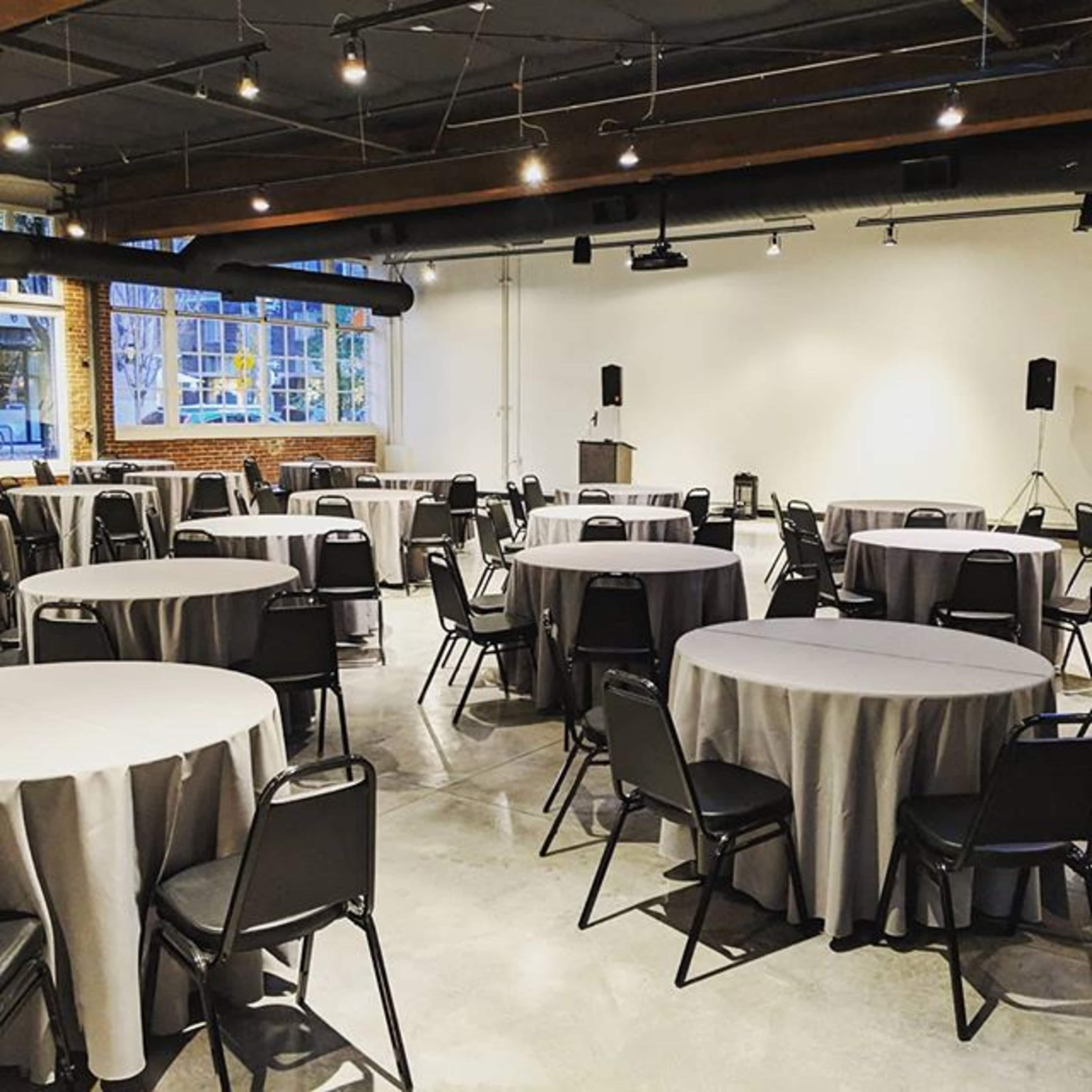 A functional event space set up with several round tables covered with gray tablecloths and black chairs arranged around them.