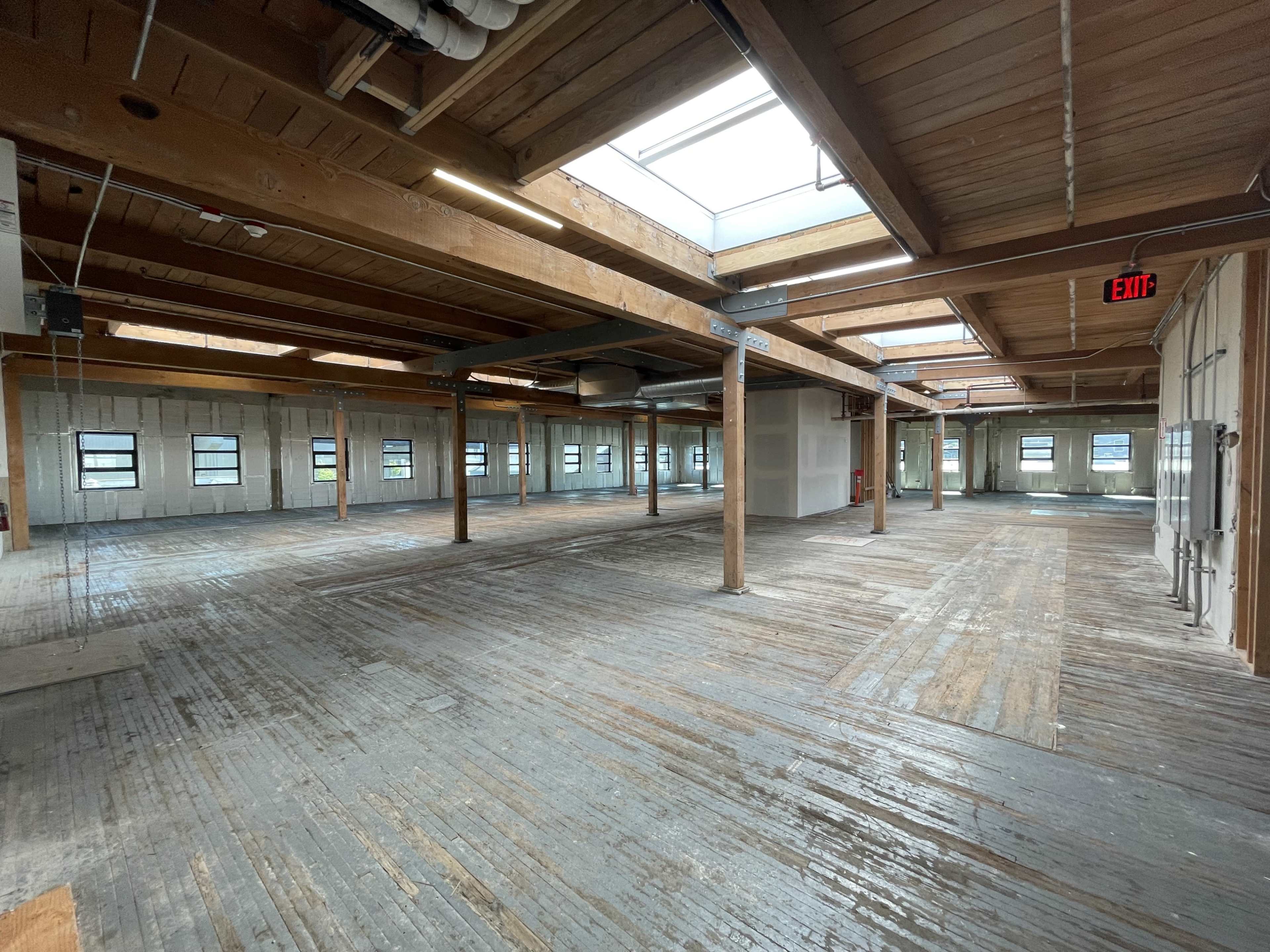 The image shows a spacious, vacant interior of a building with wooden floors and exposed beams, illuminated by natural light coming through skylights.