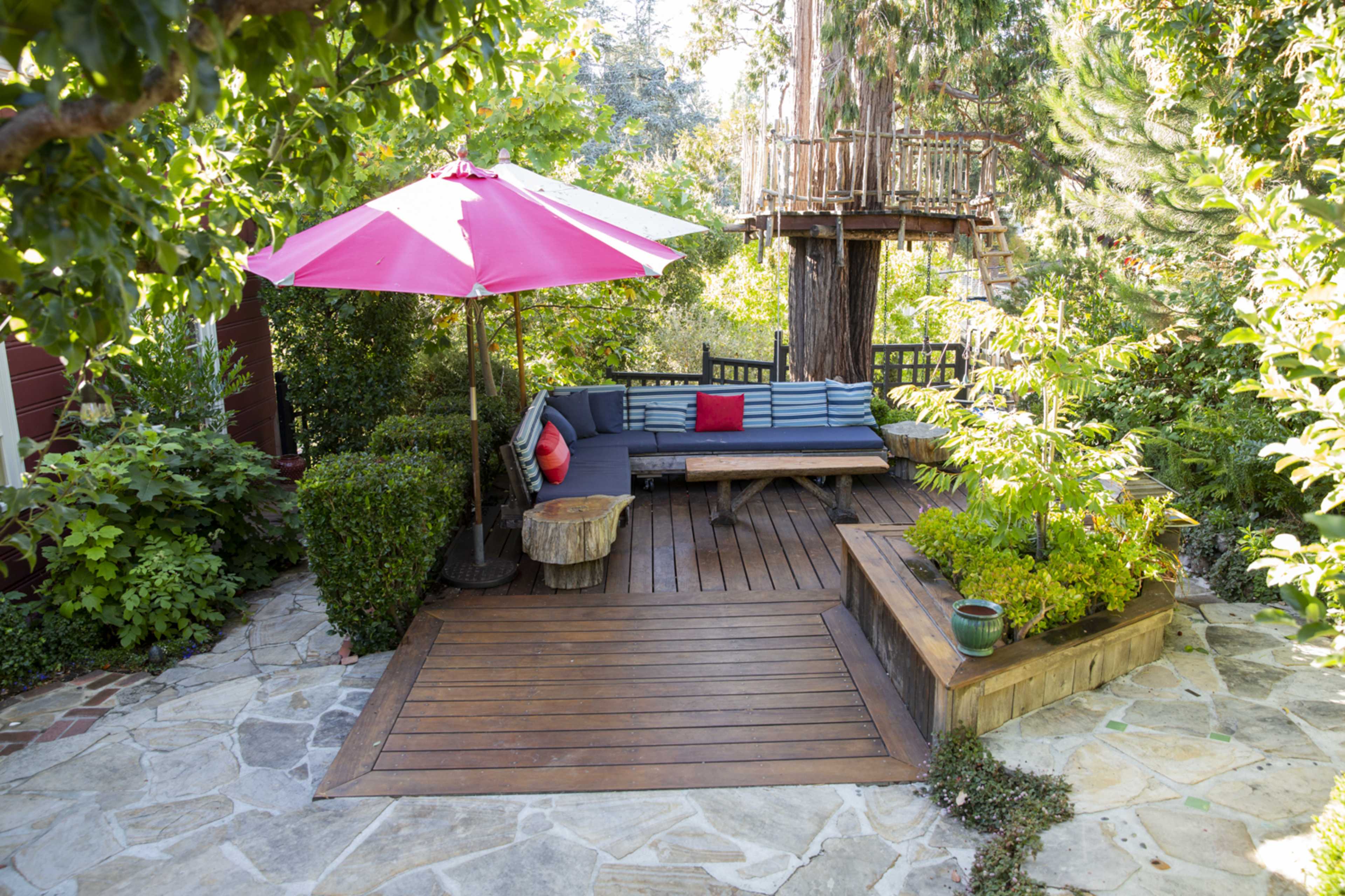 A wooden patio with a seating area, a pink umbrella, and a tree house surrounded by greenery and stone pathways.