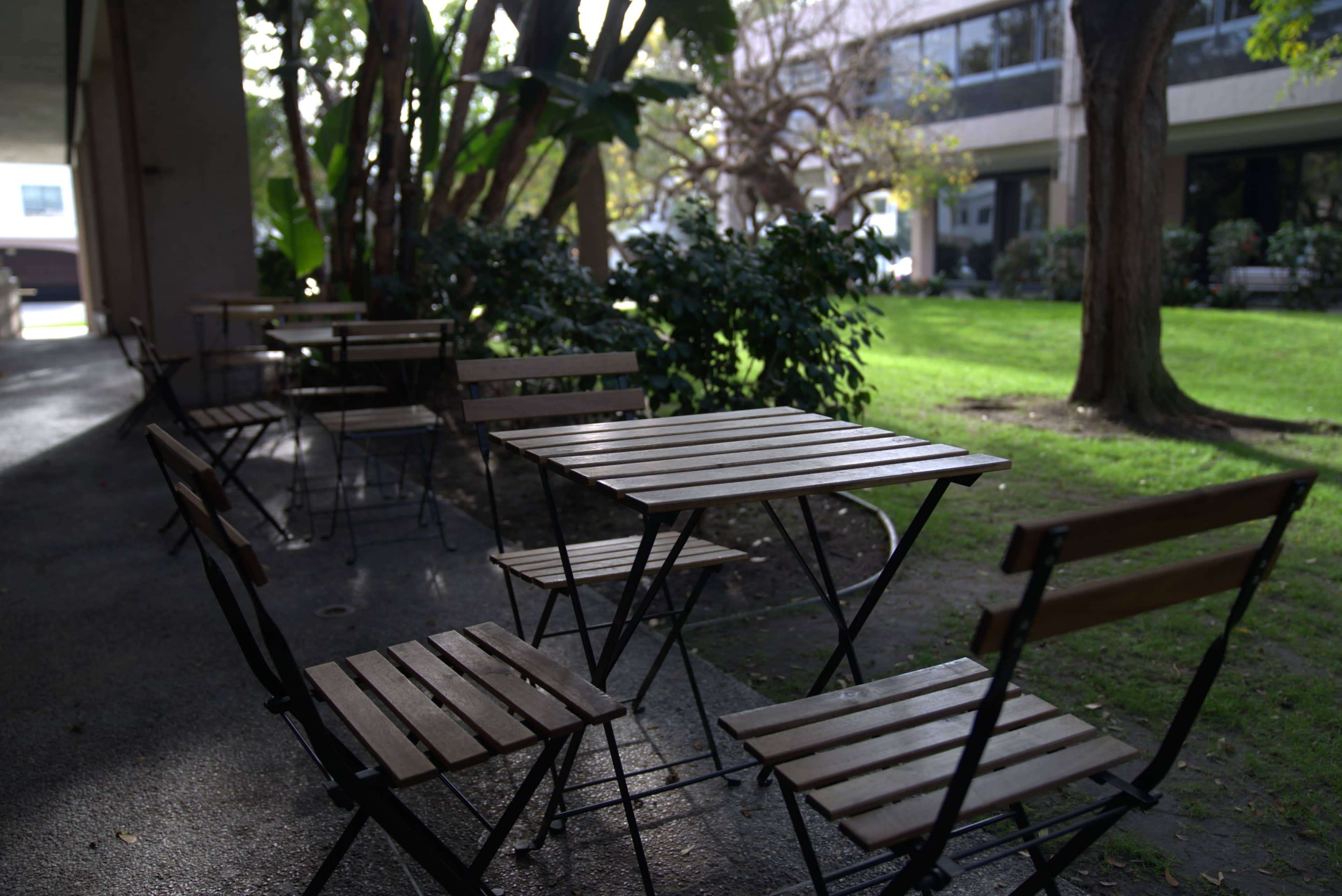 A row of wooden tables and chairs is arranged along a shaded walkway beside a grassy area and trees.