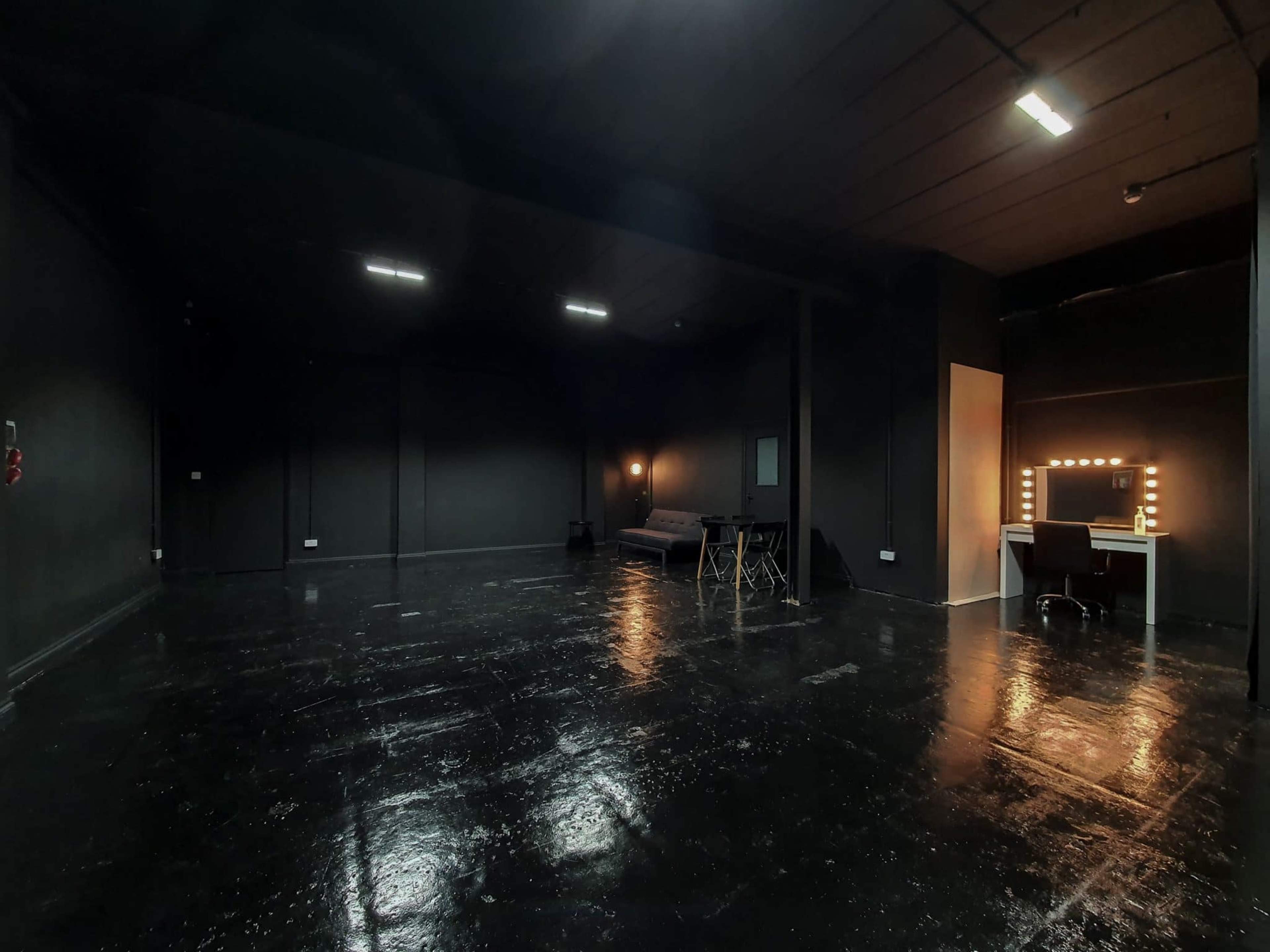 A spacious, dimly lit black room with a shiny floor, a sofa, a table with chairs, and a well-lit vanity mirror against the wall.