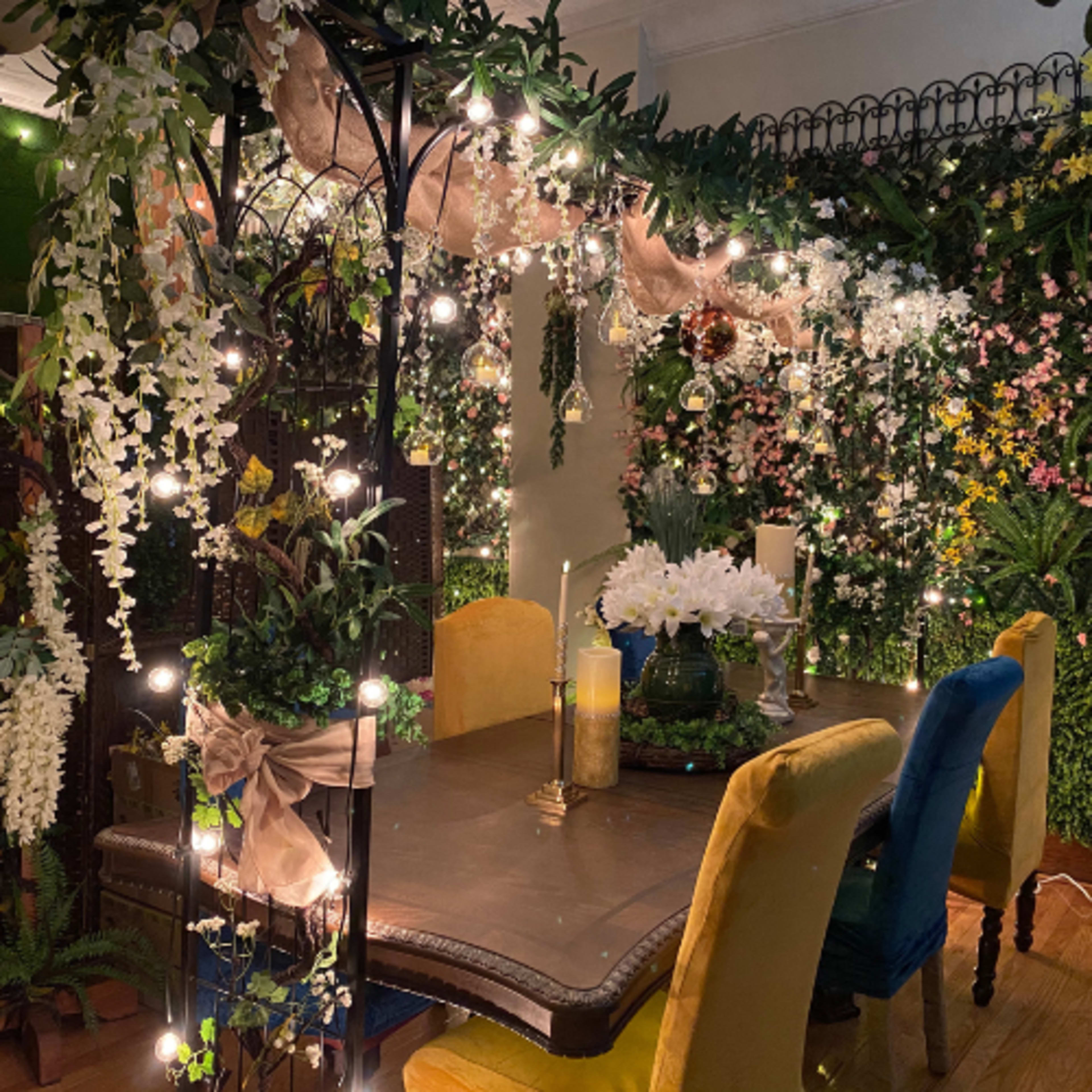 The image shows a dining area adorned with lush greenery, hanging lights, and floral decorations surrounding a wooden table set with candles and a vase of flowers.