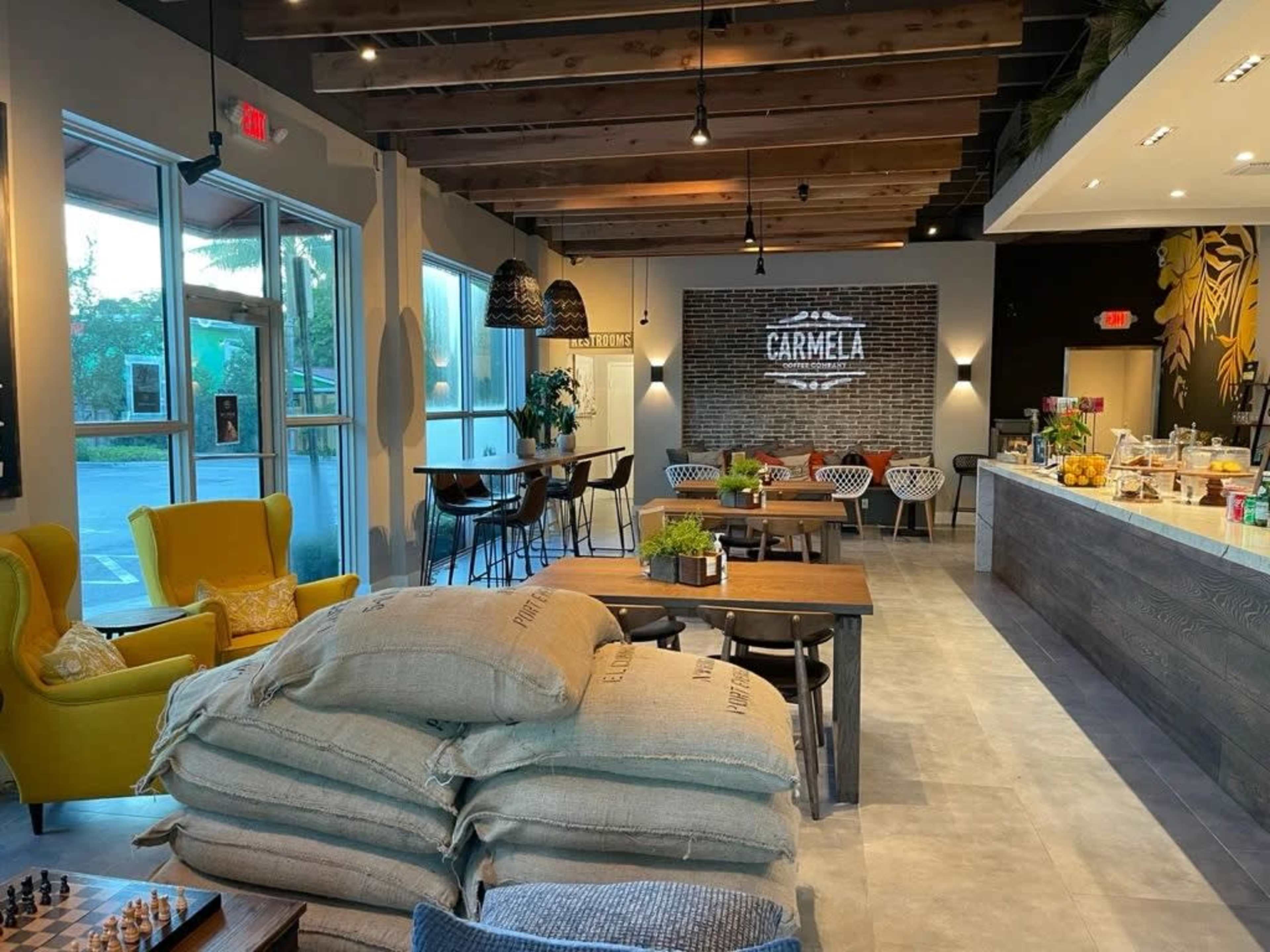 The image shows a modern café interior featuring a mix of seating options, decorative plants, and bags of coffee stored in a corner.