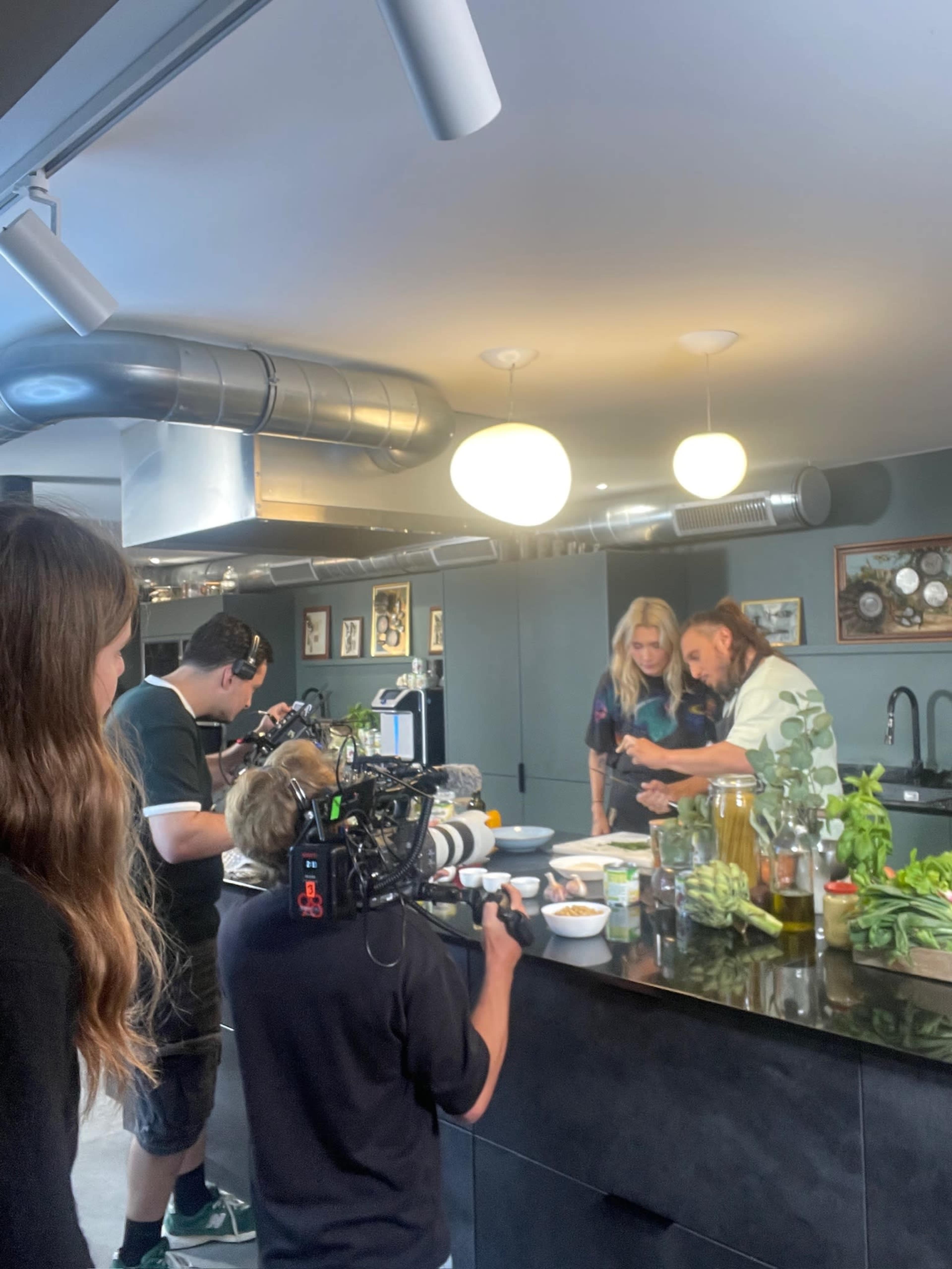 A group of individuals works in a modern kitchen, where two people are preparing food while others film the process with cameras.