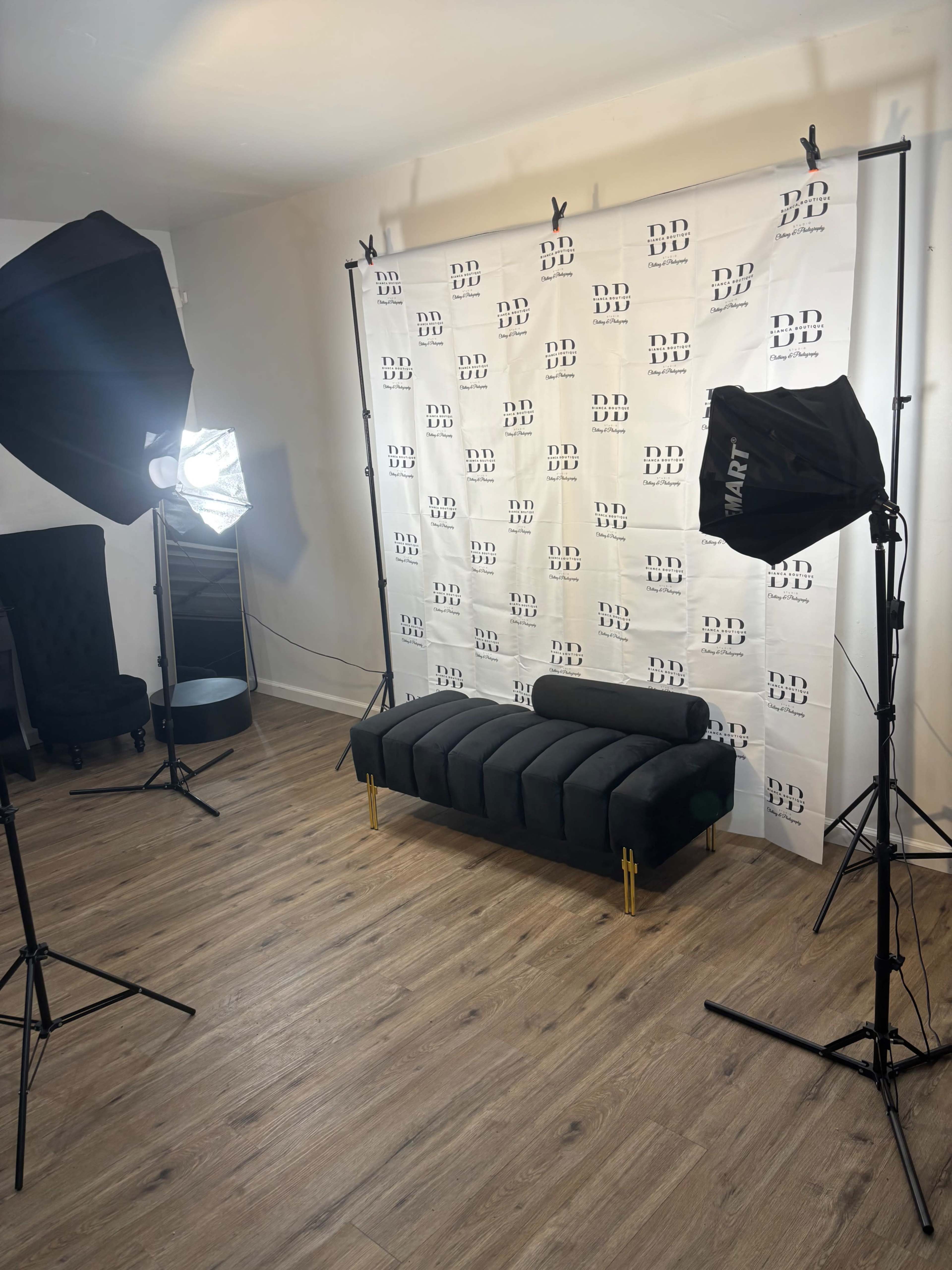 The image shows a photography studio setup featuring a black sofa in front of a backdrop with repeated text, flanked by two softbox lighting setups.