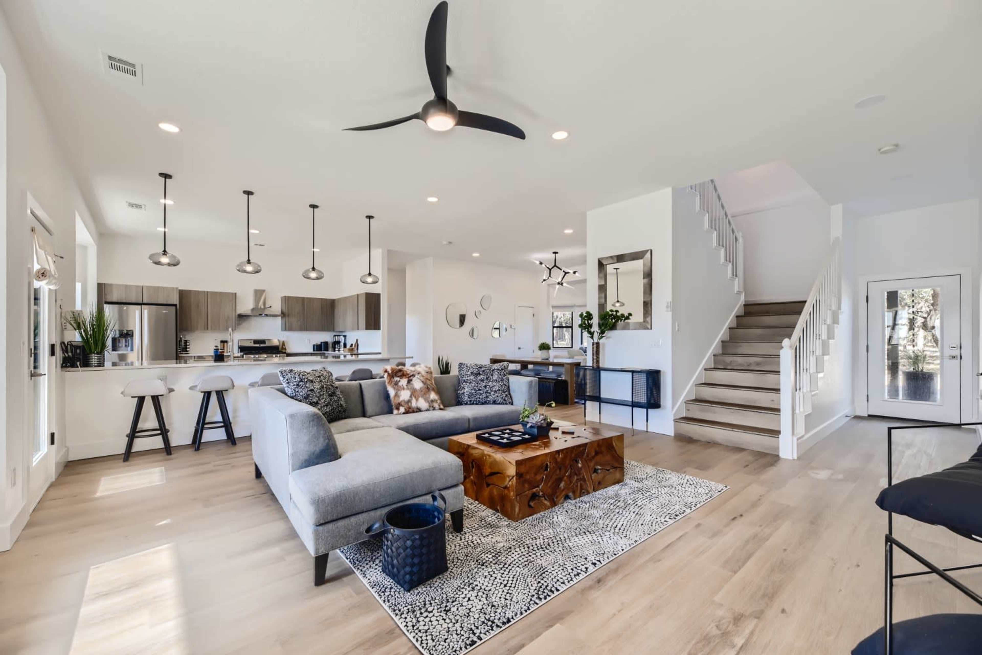 A modern open-concept living room and kitchen area featuring a sectional sofa, wooden coffee table, and a staircase leading to an upper level.