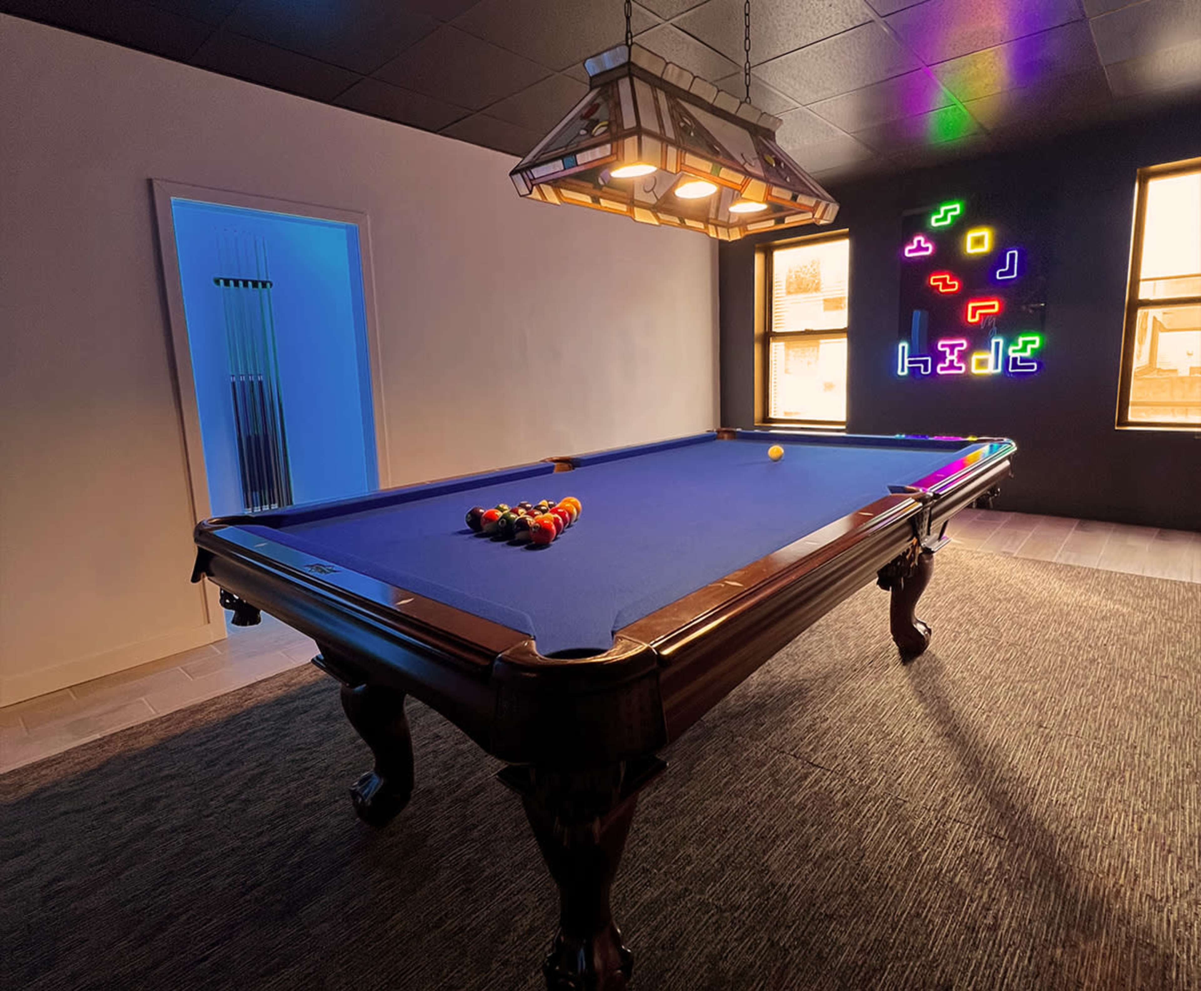 A pool table with a blue felt surface is positioned in a well-lit room featuring a neon sign on the wall and a doorway leading to another space.