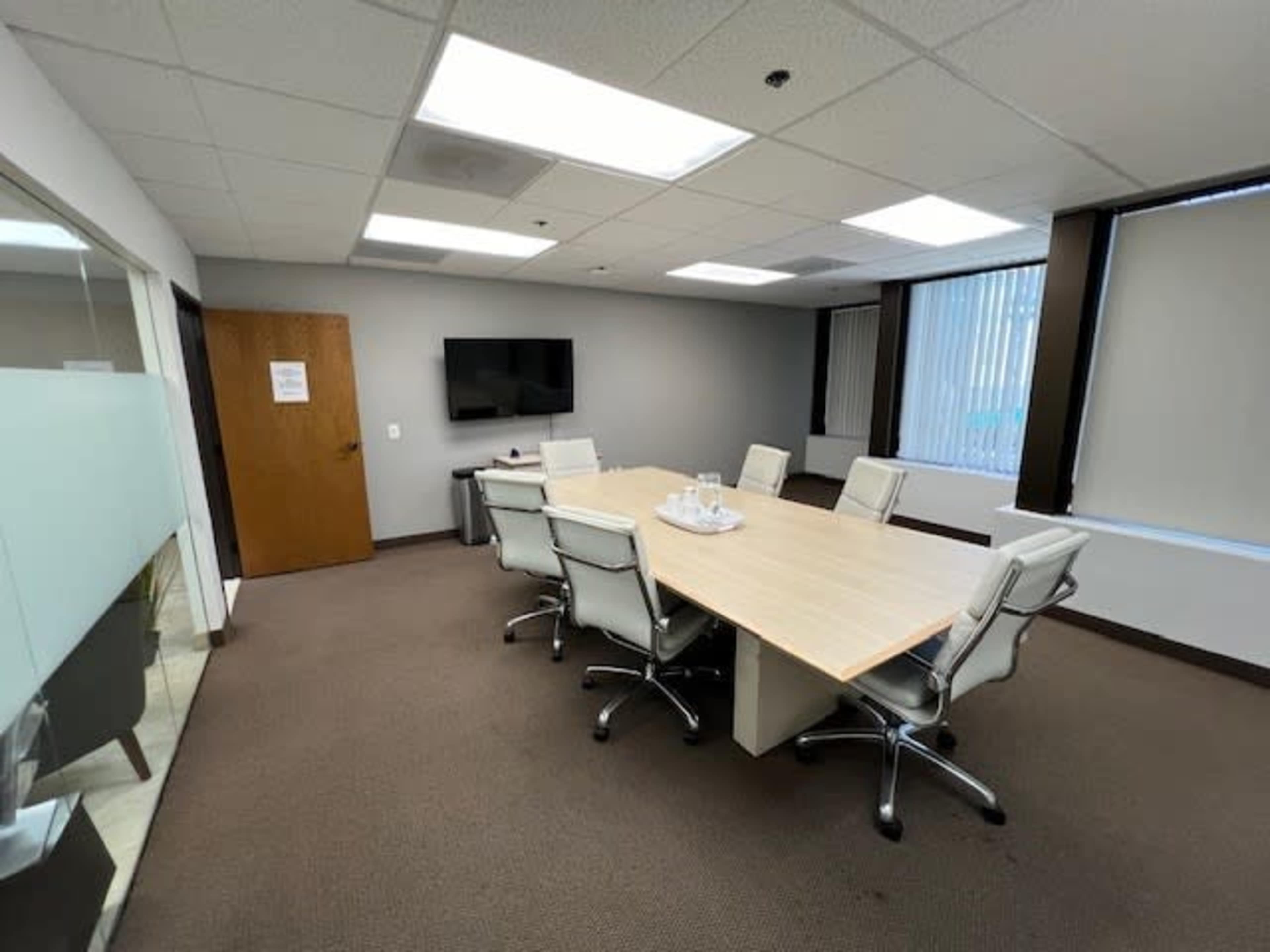 A conference room features a large wooden table surrounded by six silver office chairs, with a television mounted on the wall and windows dressed in vertical blinds.