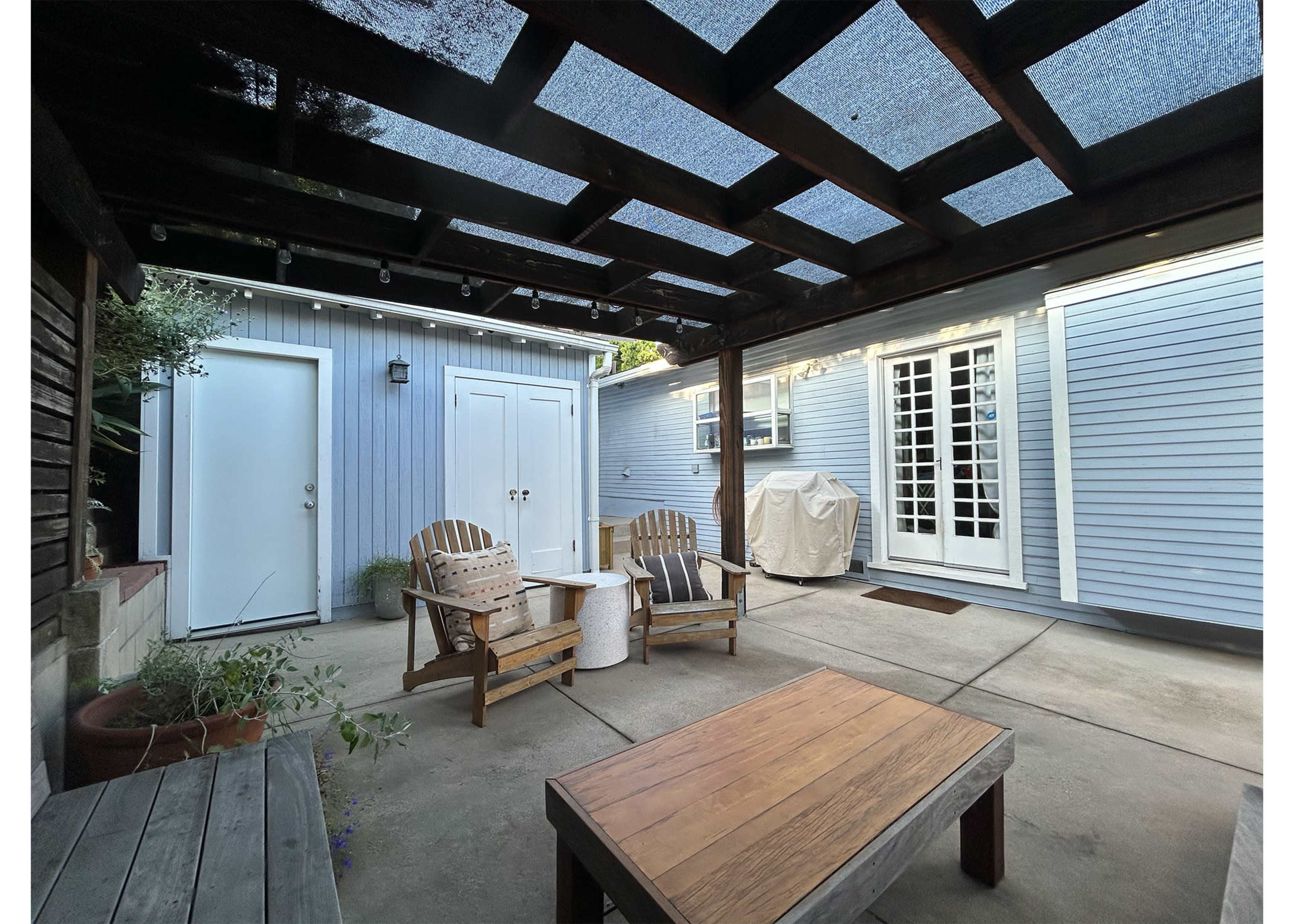 The image shows a patio area with two wooden chairs, a small table, and a door leading to a building in the background, enclosed by walls and a slatted roof.