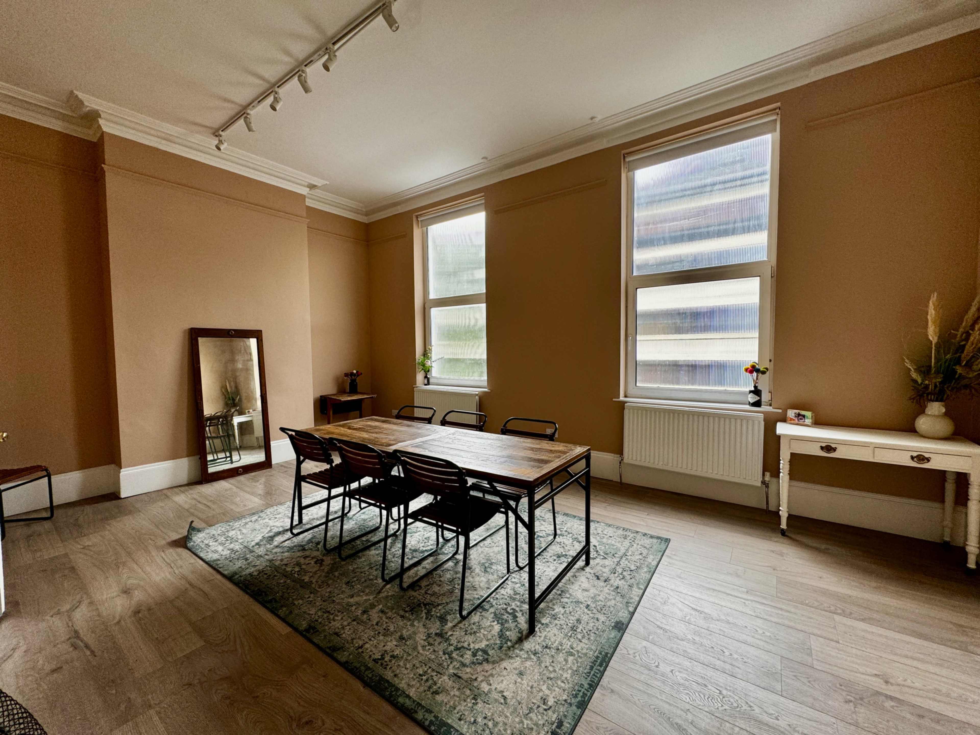 The image shows a spacious room with a wooden table surrounded by chairs, a mirror against one wall, and large windows letting in natural light.