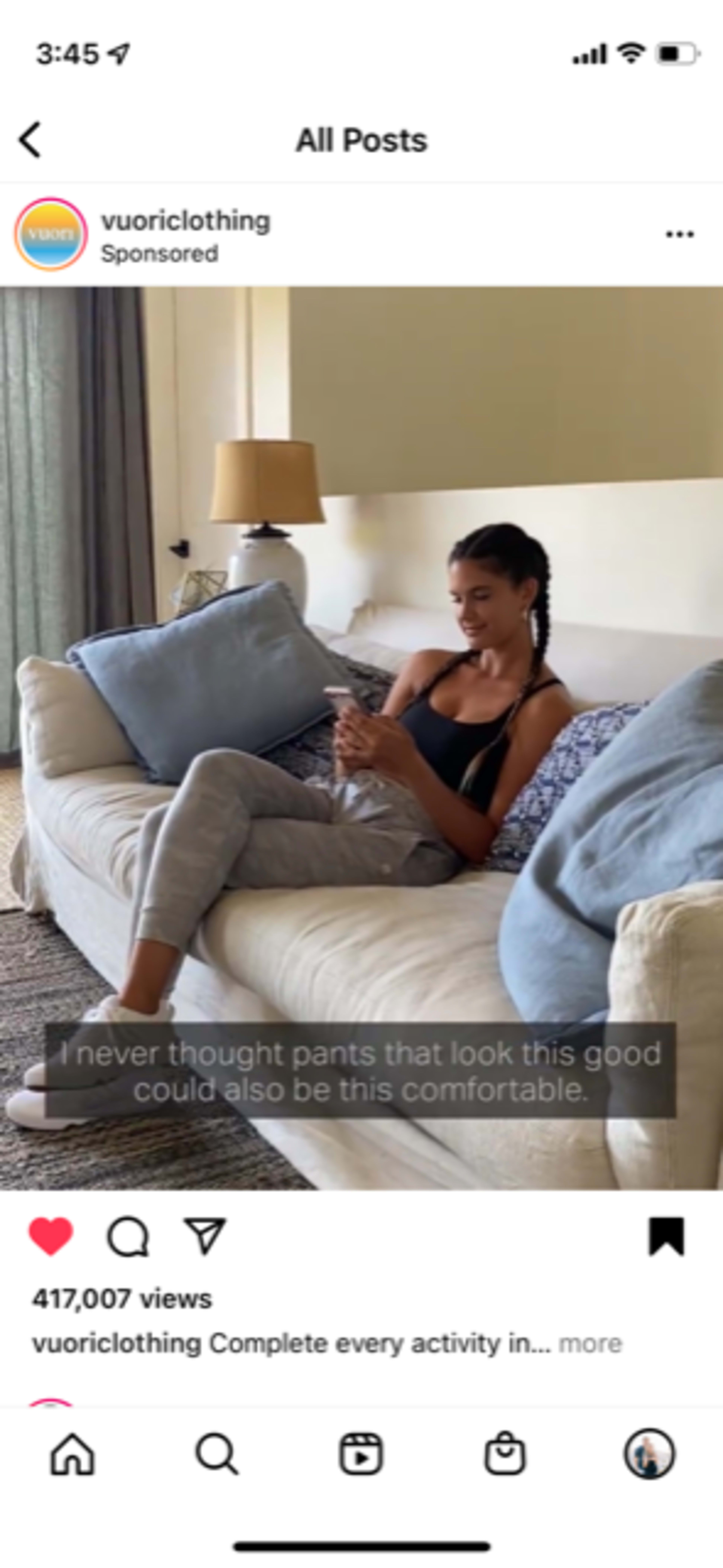 A woman sits on a couch in a living room, looking at her phone while wearing comfortable clothing.