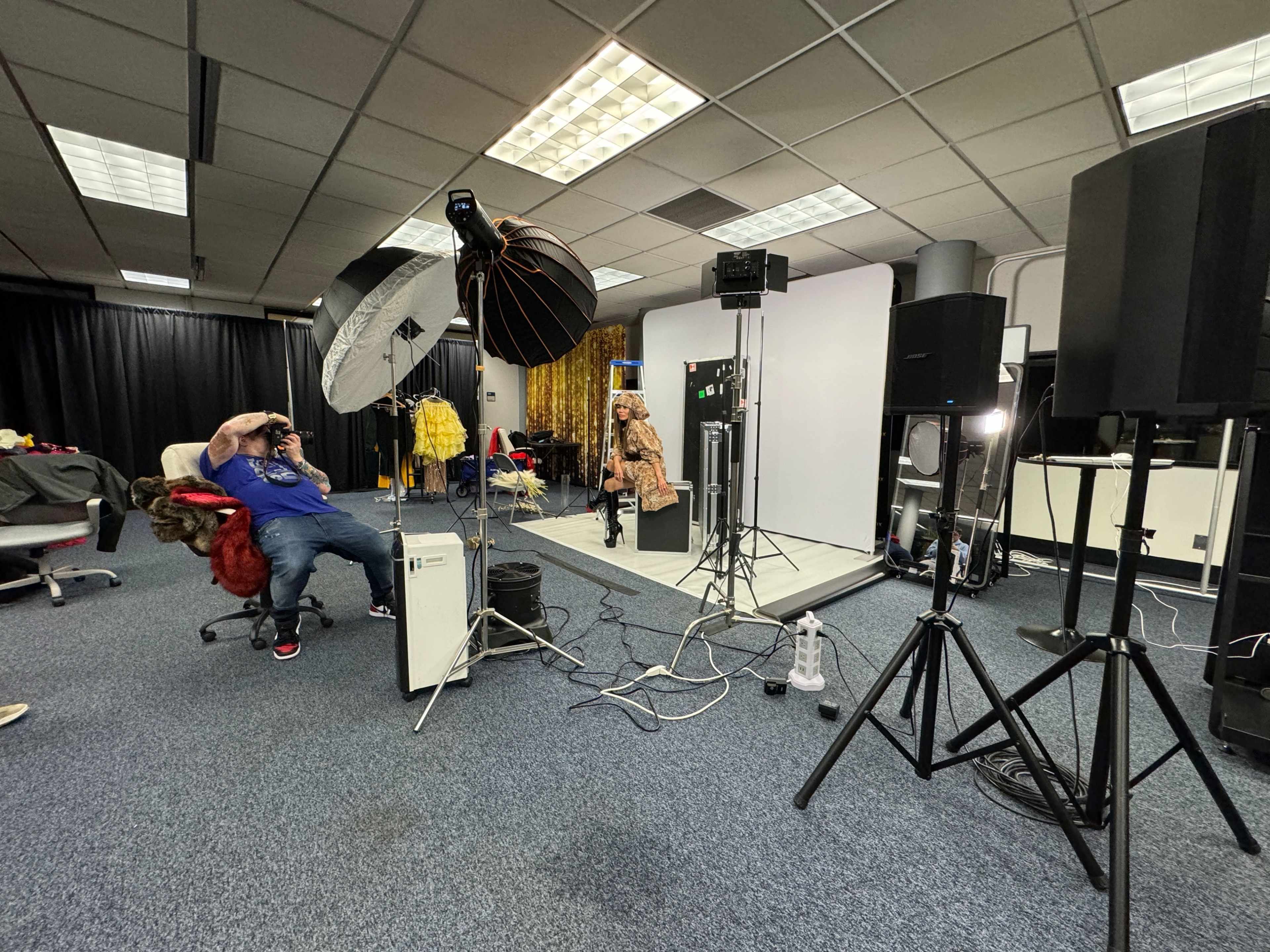 The image shows a photography studio setup with a model posing against a backdrop, surrounded by multiple lights, camera equipment, and a photographer in a chair.