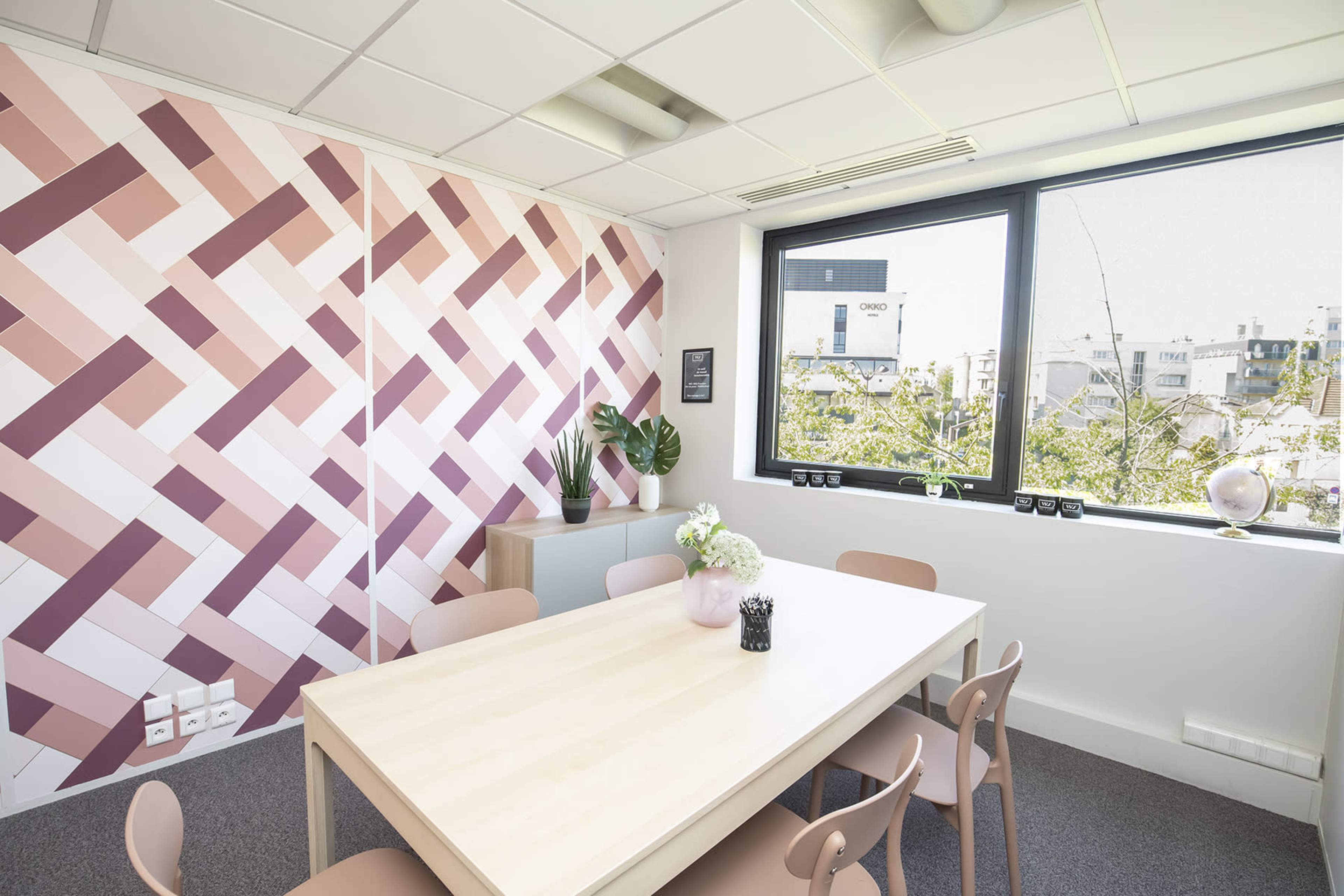 A modern meeting room features a light wooden table surrounded by pastel chairs, with a patterned pink wall and large windows overlooking a cityscape.