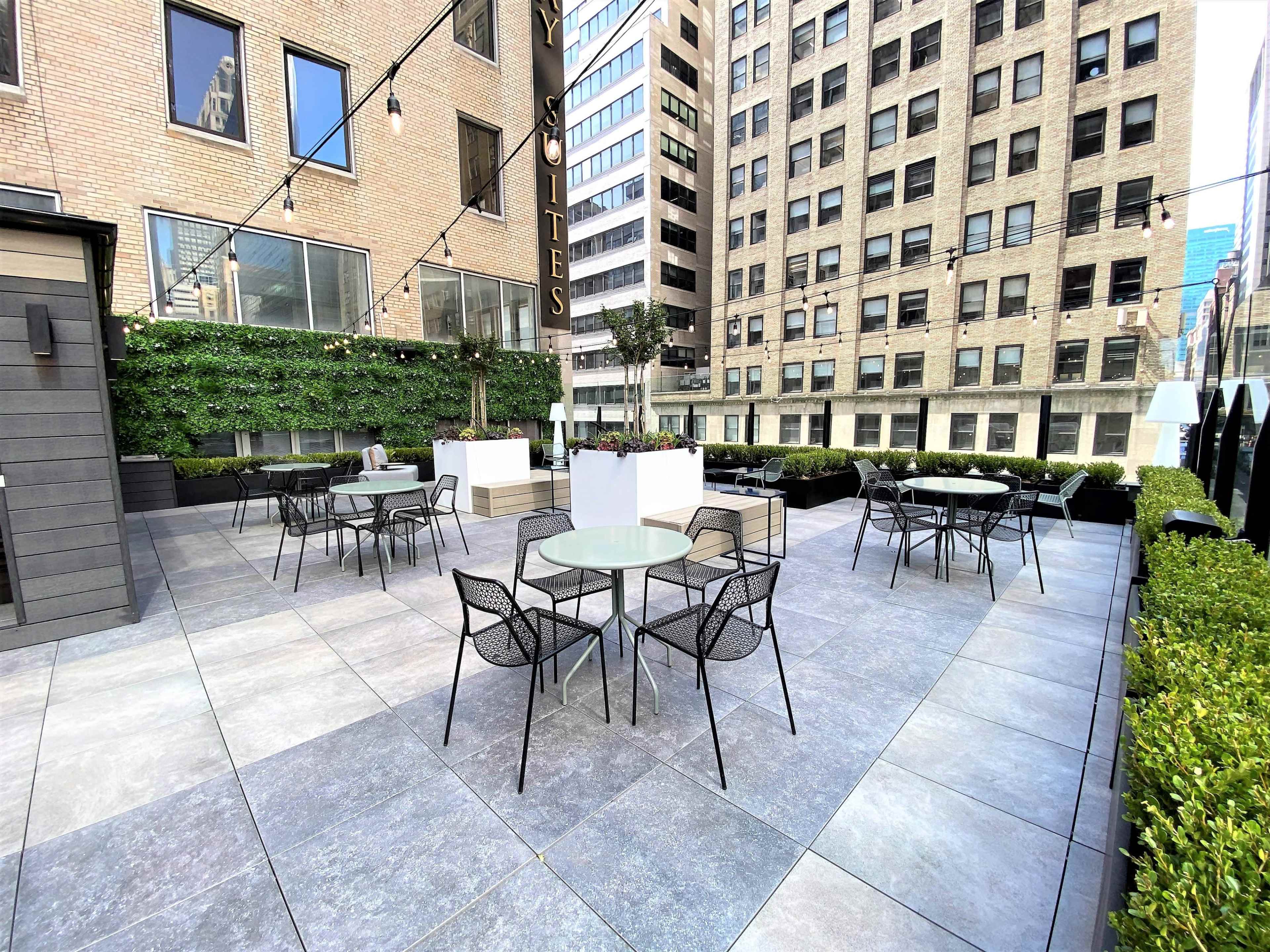 The image shows a rooftop terrace with several glass-top tables and black chairs surrounded by green planters and urban buildings.