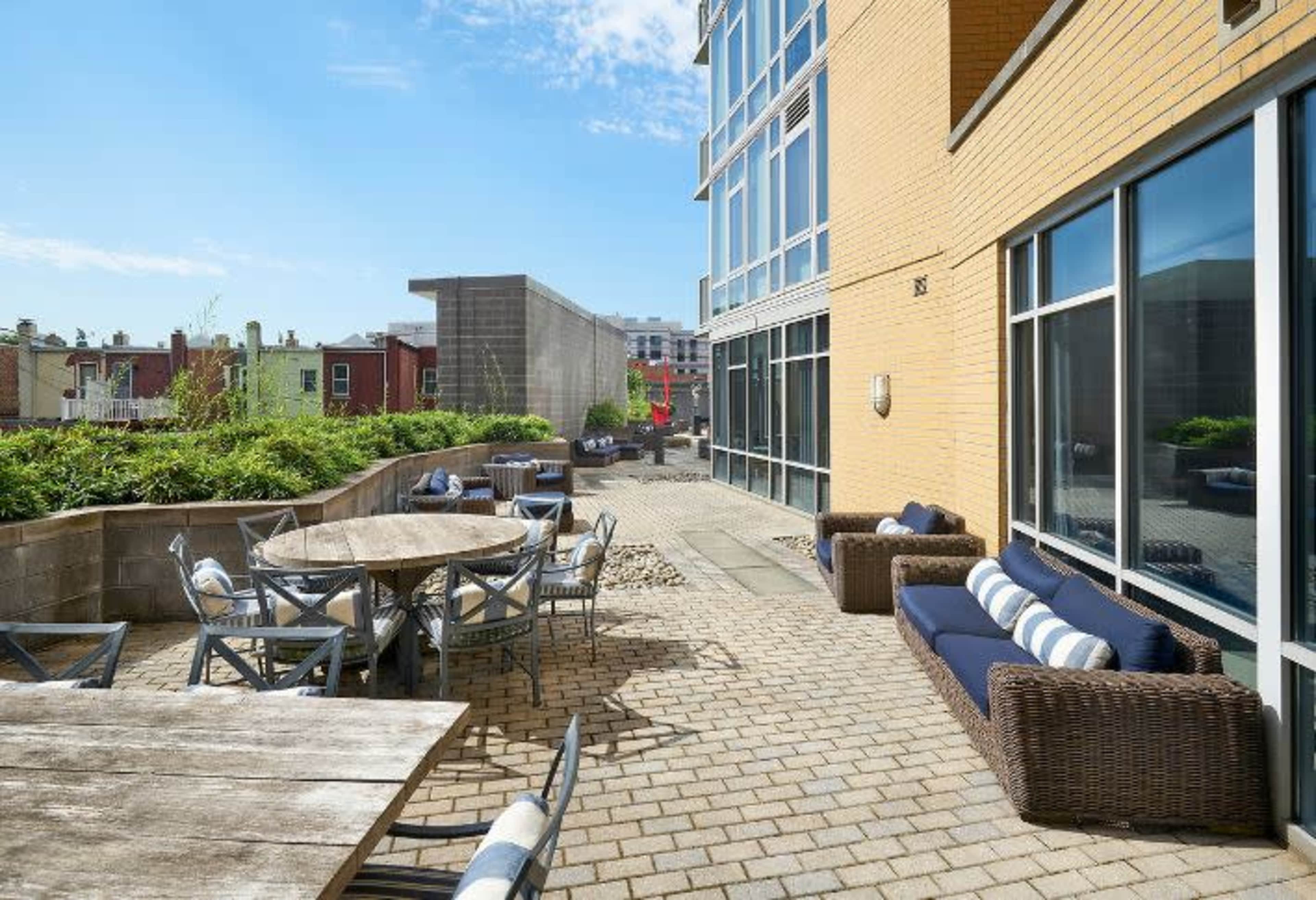The image shows a spacious outdoor patio area with wooden tables, metal chairs, and modern seating, surrounded by greenery and buildings in the background.