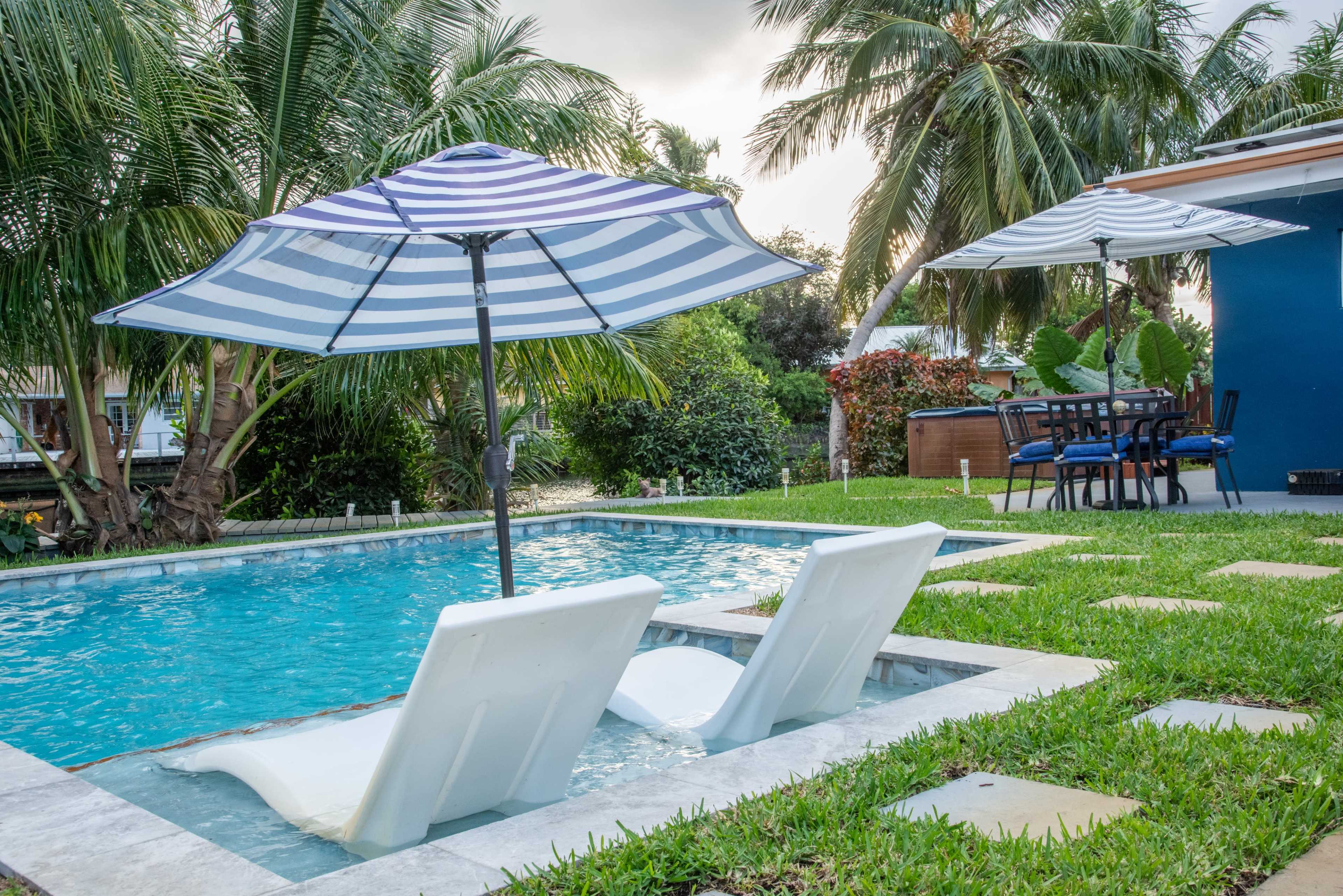 The image shows a poolside area with two white lounge chairs under striped umbrellas, surrounded by green grass and palm trees, with a dining table and chairs nearby.