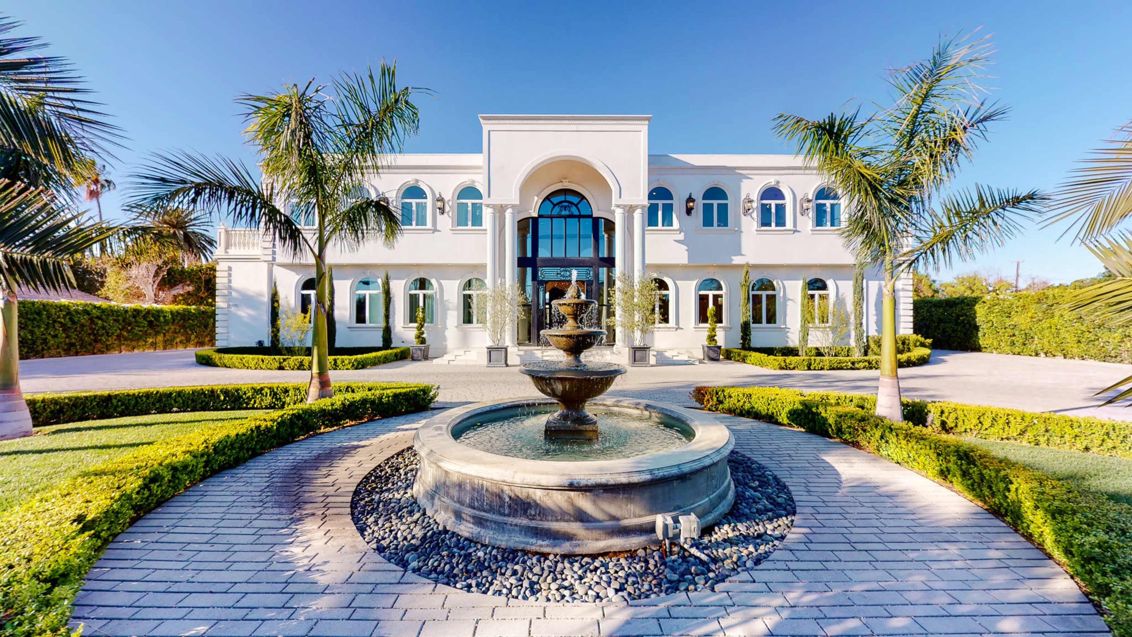 A large, two-story white villa with a central fountain and palm trees in the foreground.