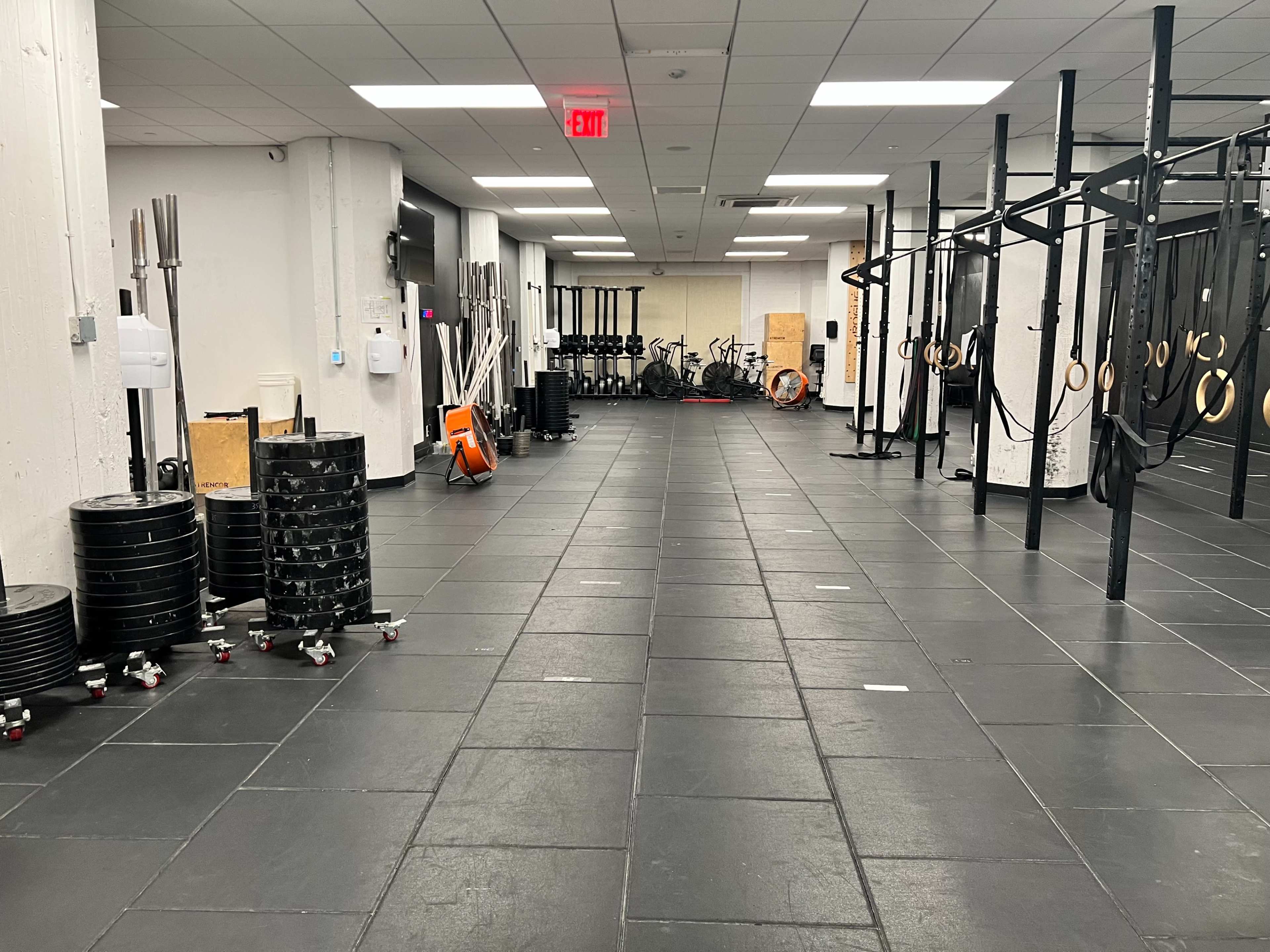 An empty gym space with black rubber flooring, weight plates on wheels, exercise equipment, and a row of gymnastic rings hanging from a frame.