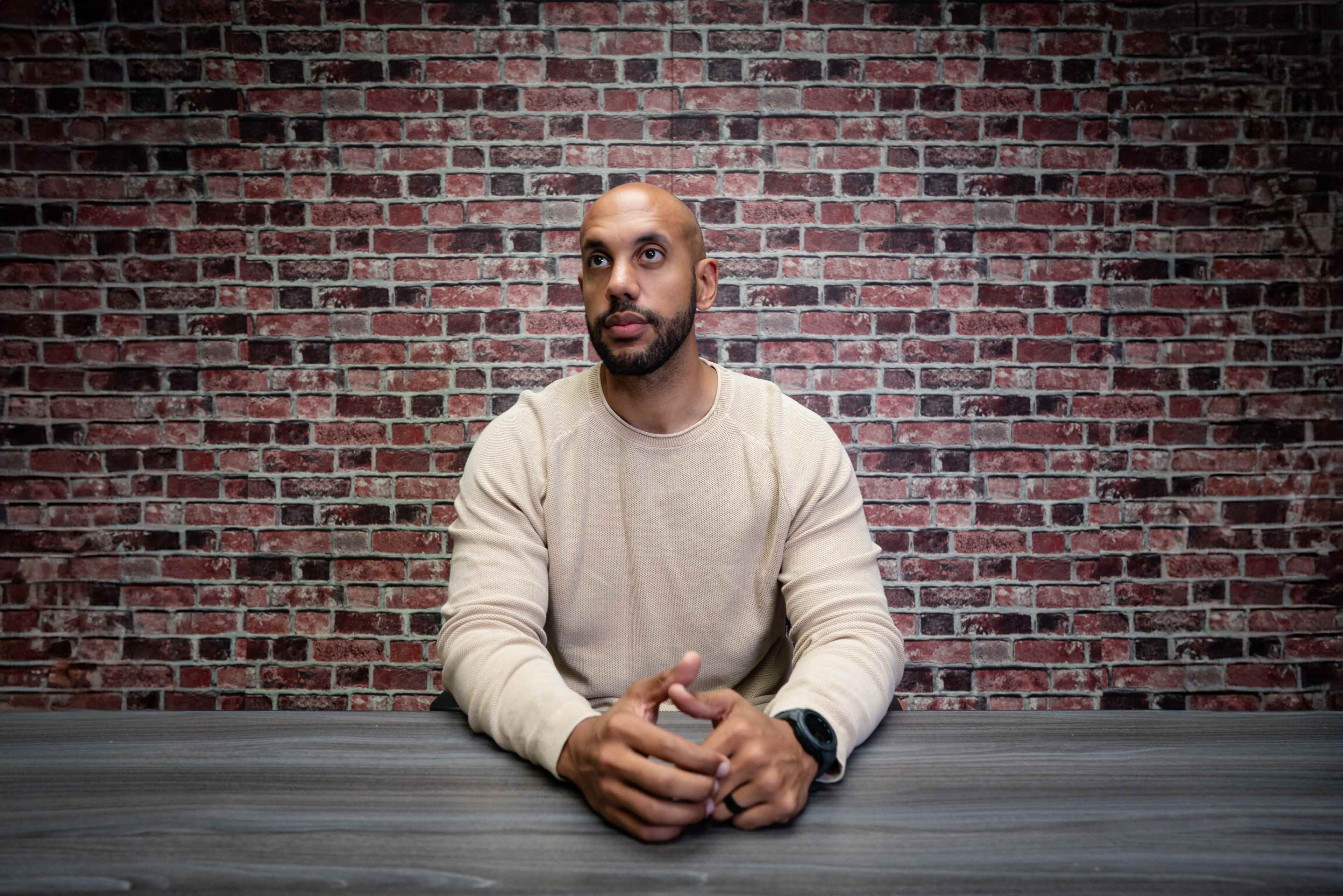 A man with a short beard sits at a wooden table in front of a brick wall.