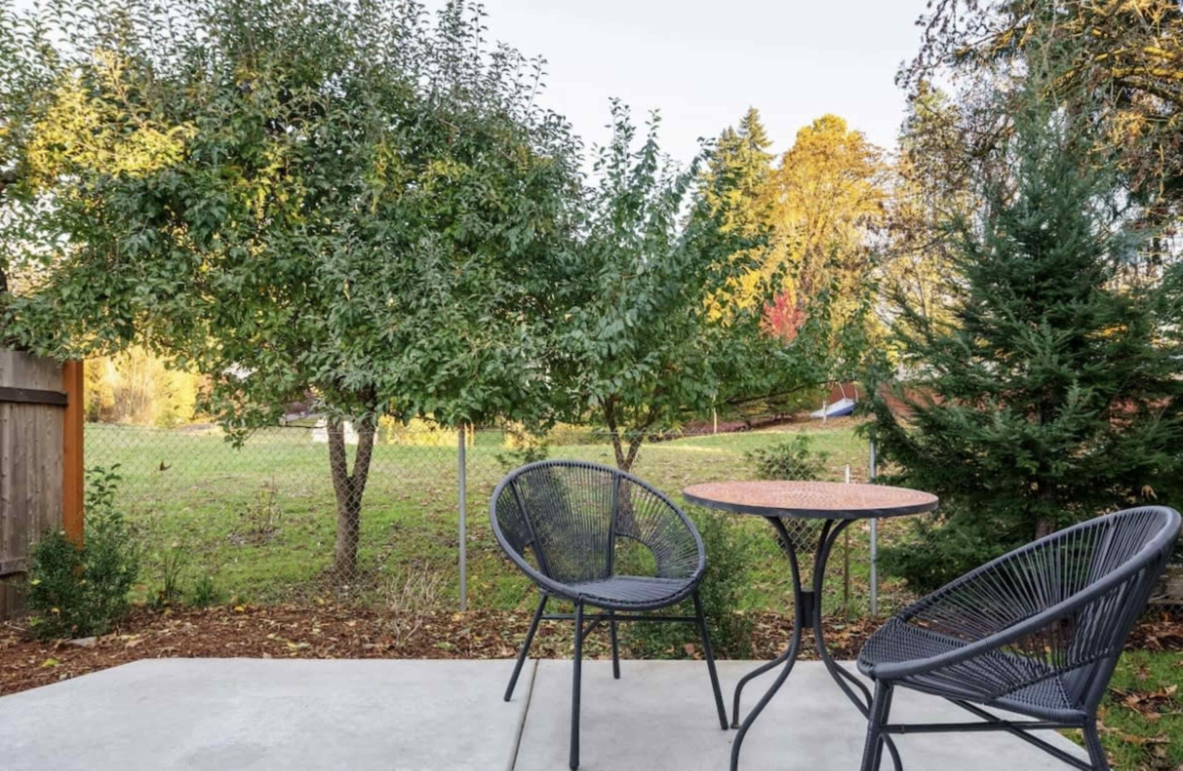 A small patio features two chairs and a round table, with a backdrop of trees and an open grassy area beyond a fence.