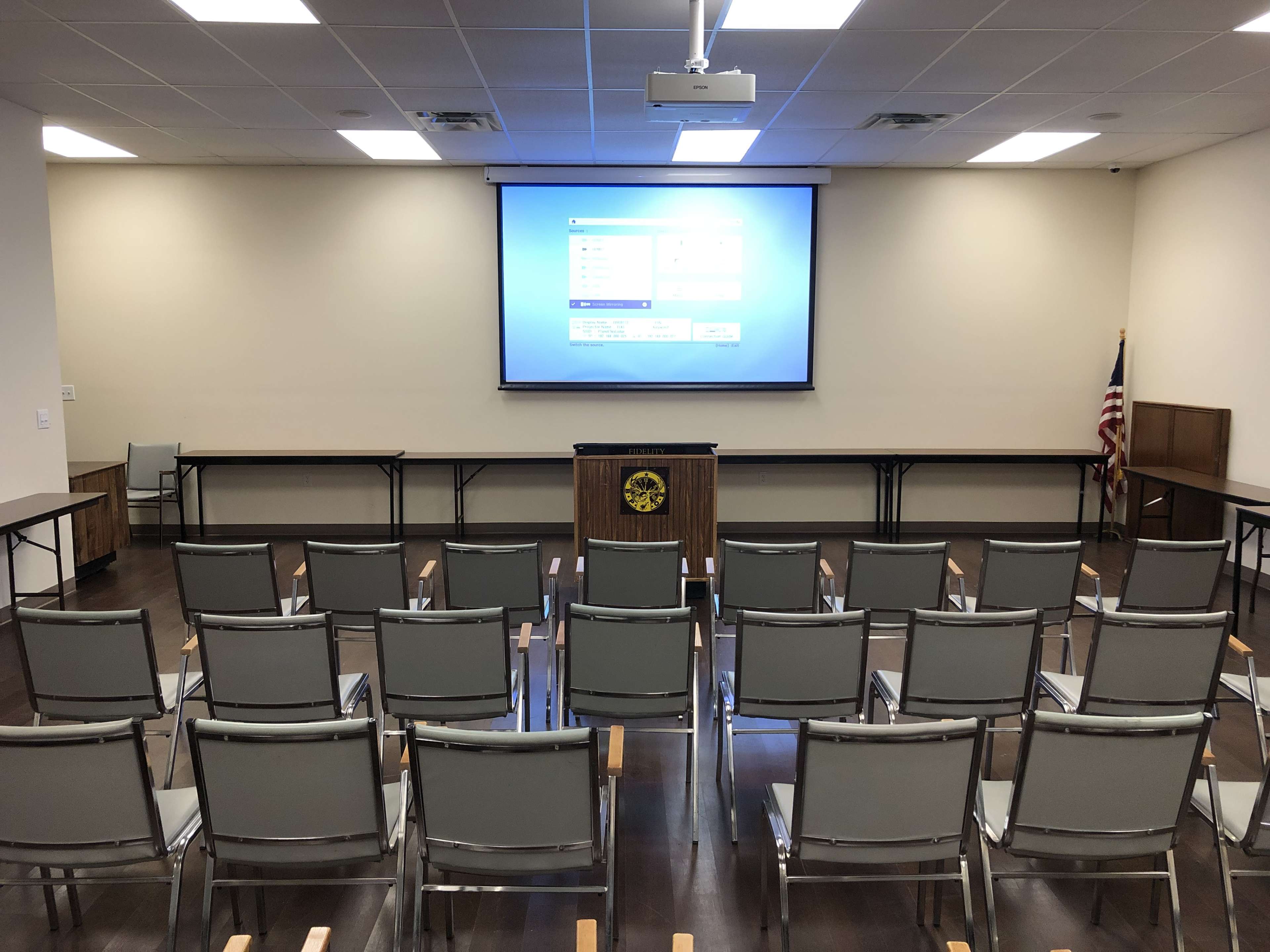 A meeting room is arranged with rows of gray chairs facing a large projection screen at the front.