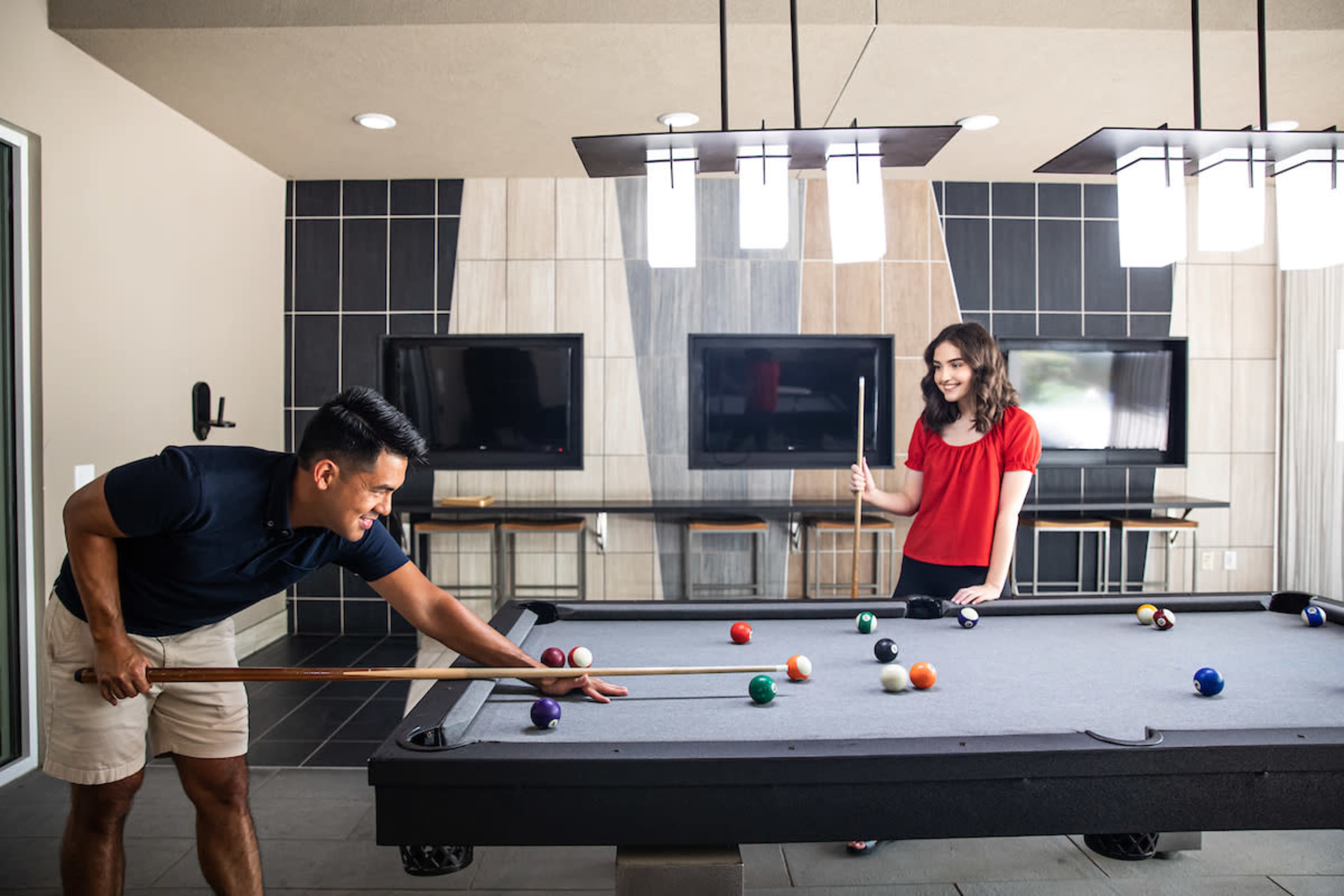 Two people are playing pool in a modern game room with a large table and multiple flat-screen TVs in the background.