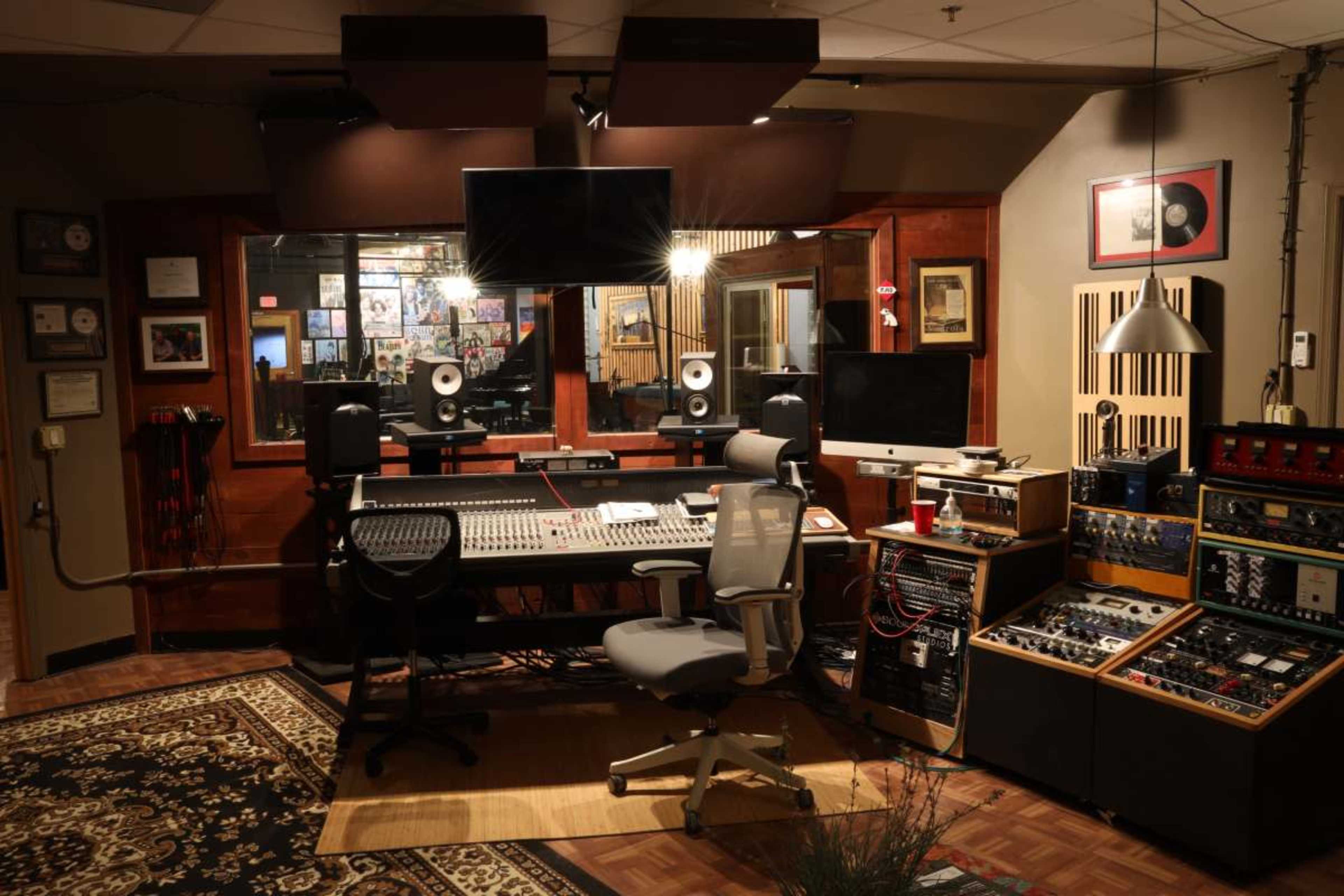 The image shows a well-equipped recording studio featuring a mixing desk, monitors, and various audio equipment, surrounded by soundproofing materials and decorative items.