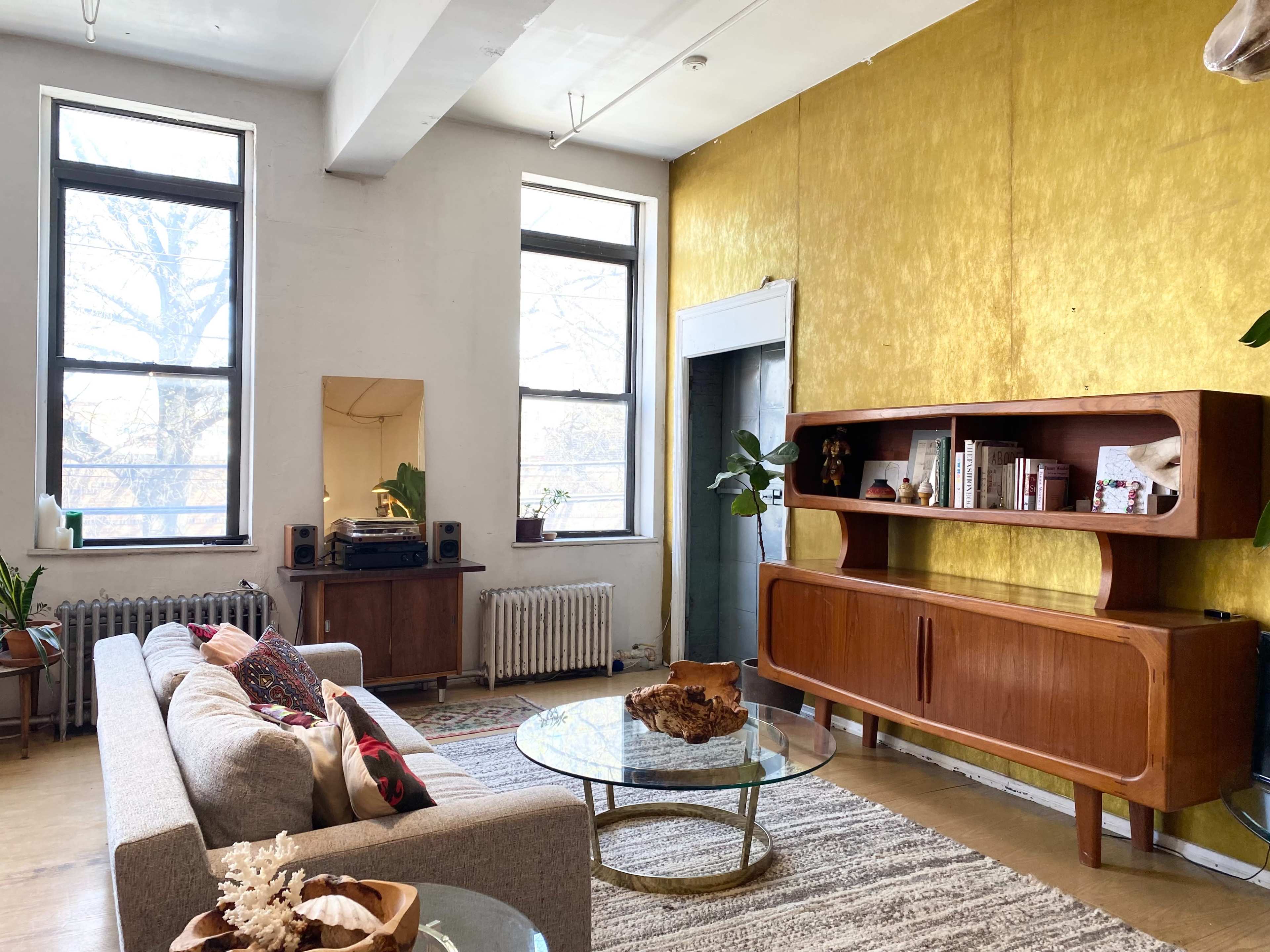 A living room with large windows, a stylish gray sofa, a wooden sideboard, and a round glass coffee table placed on a patterned rug.