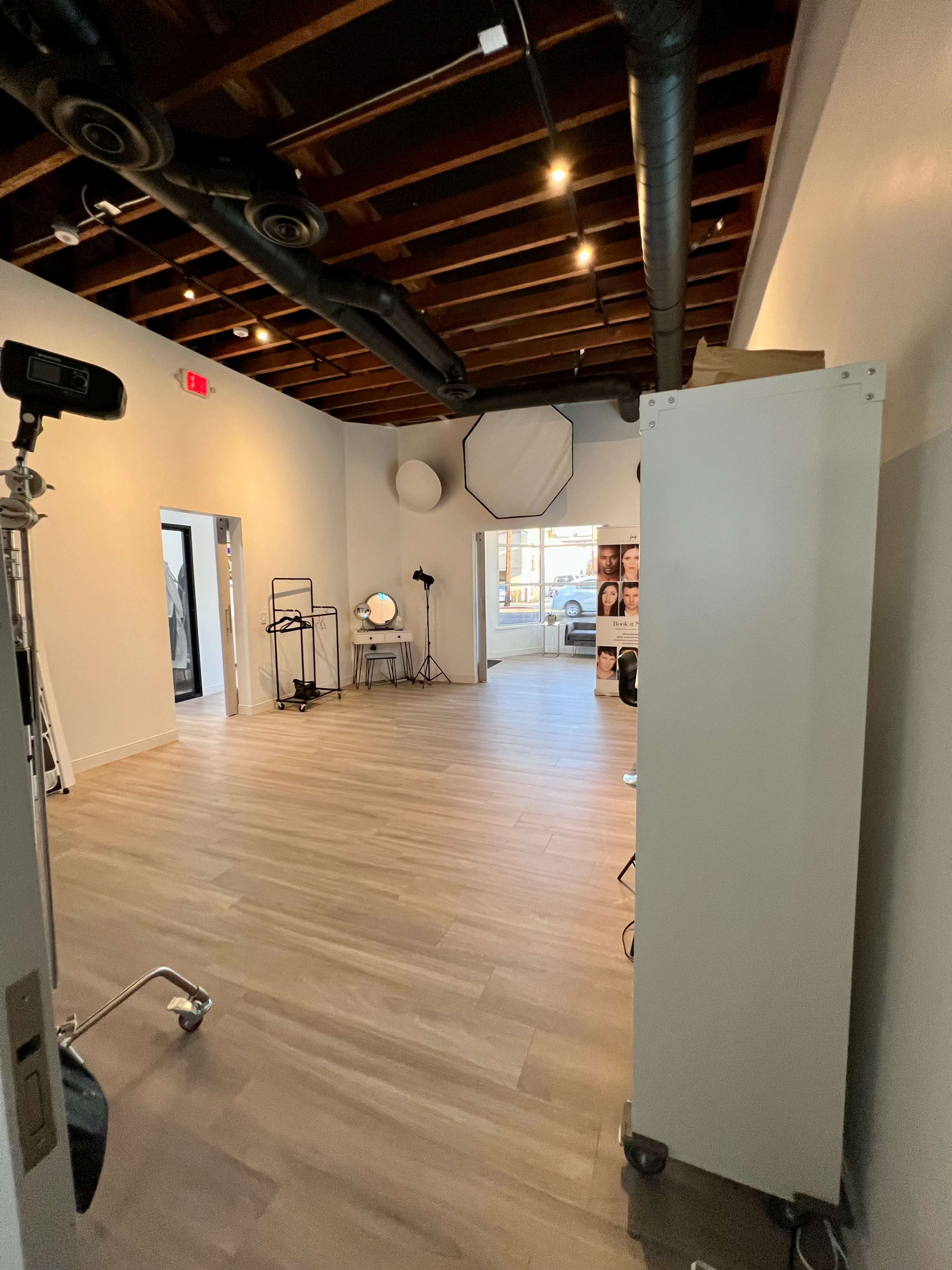 A spacious, well-lit studio with wooden flooring, large windows, and various photography equipment arranged throughout the room.