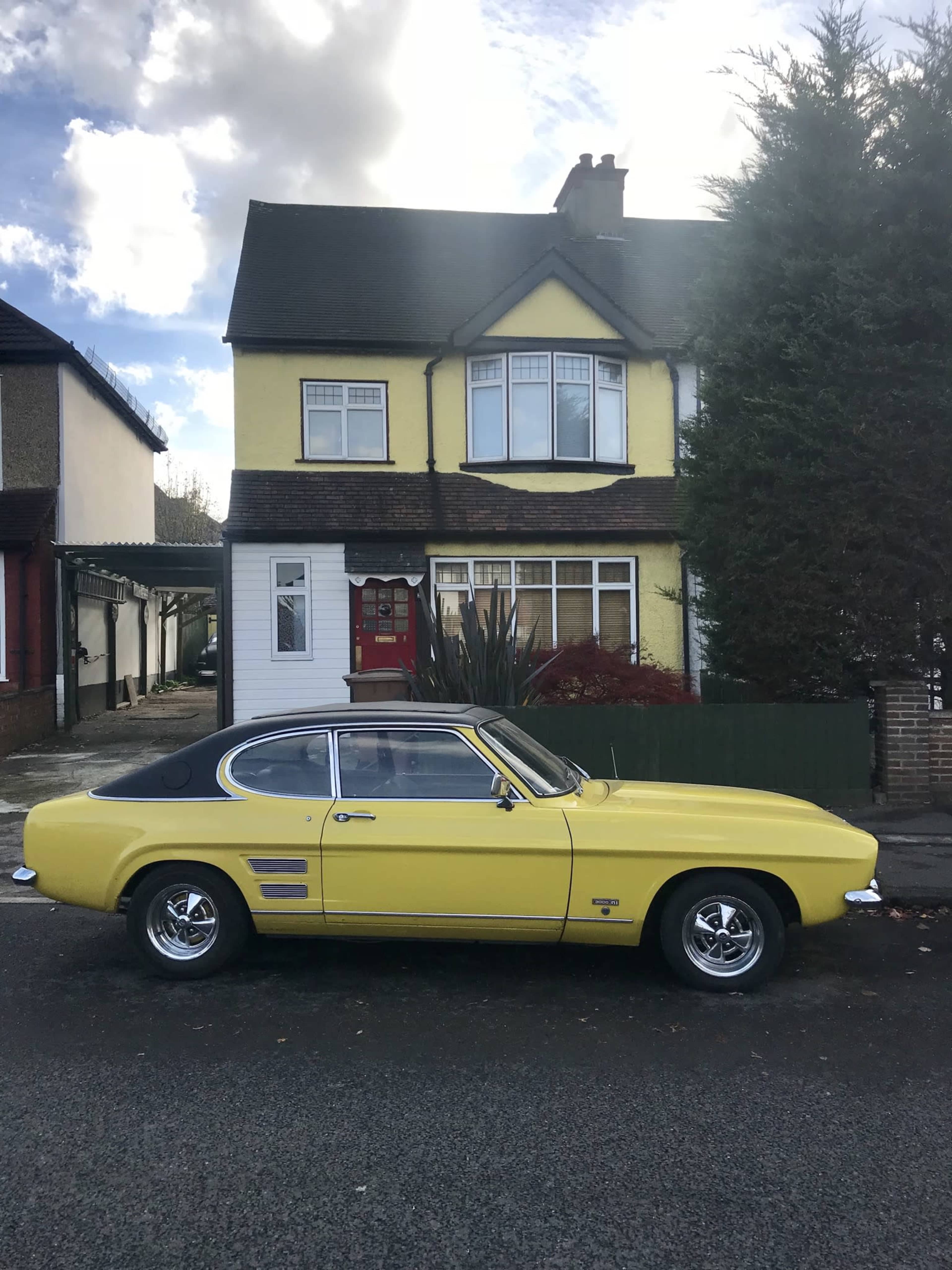 A yellow classic car is parked on the street in front of a two-story house with a distinctive yellow exterior and large windows.