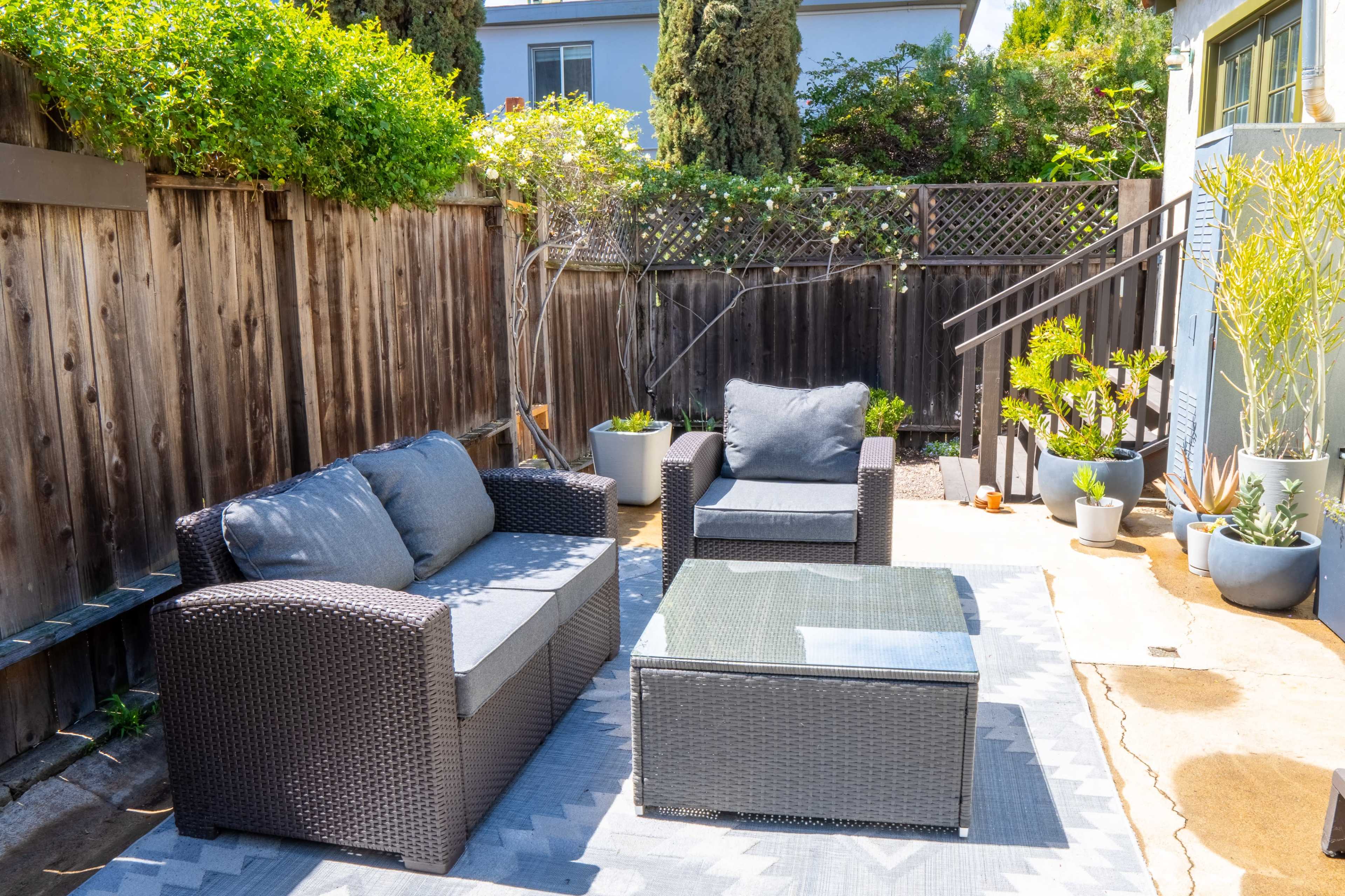 A patio area featuring a wicker seating arrangement with two chairs and a sofa, accompanied by a glass-topped coffee table and several potted plants.