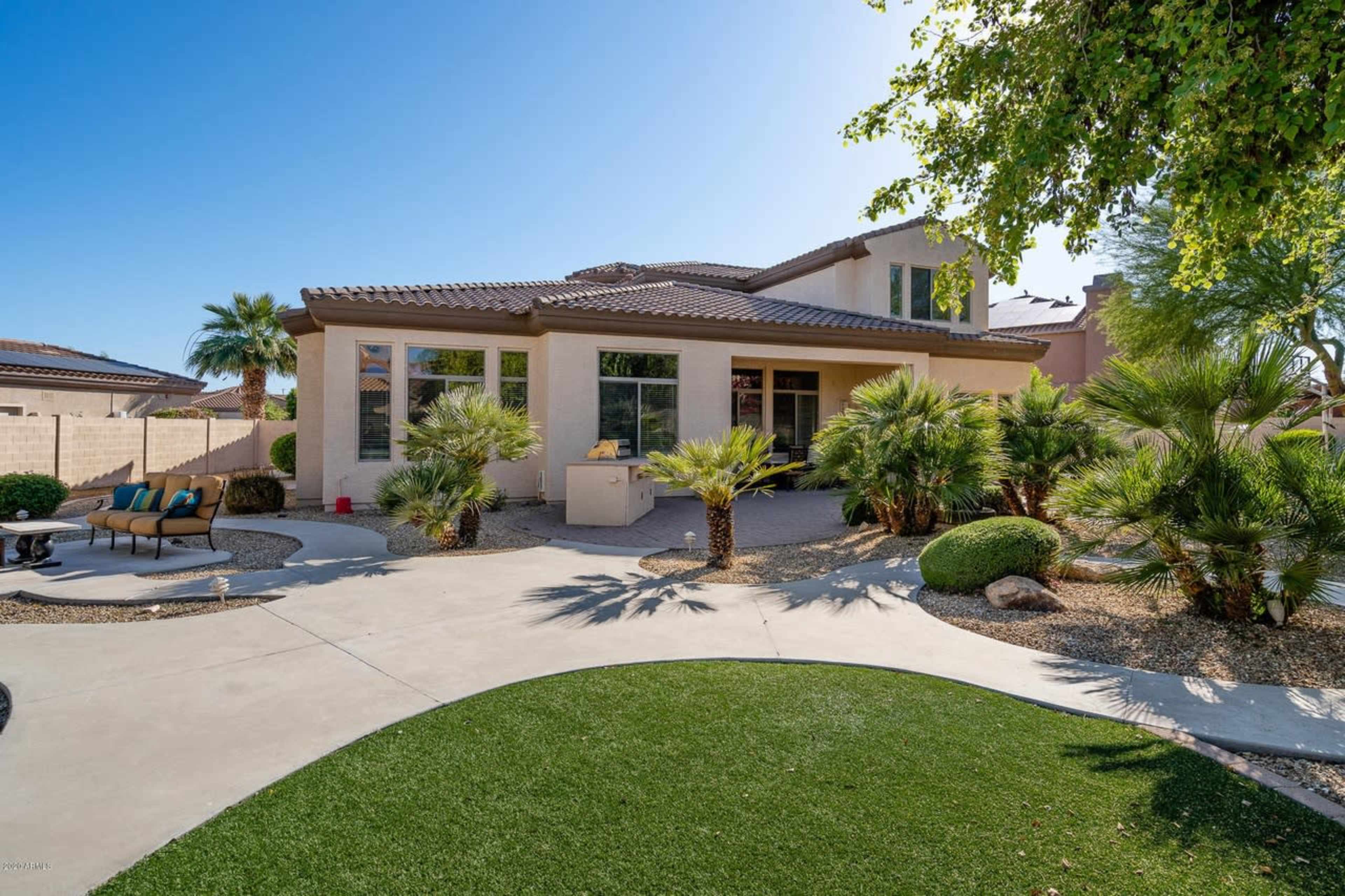 A landscaped backyard features a patio area with seating, surrounded by palm trees and greenery, leading to a two-story house.