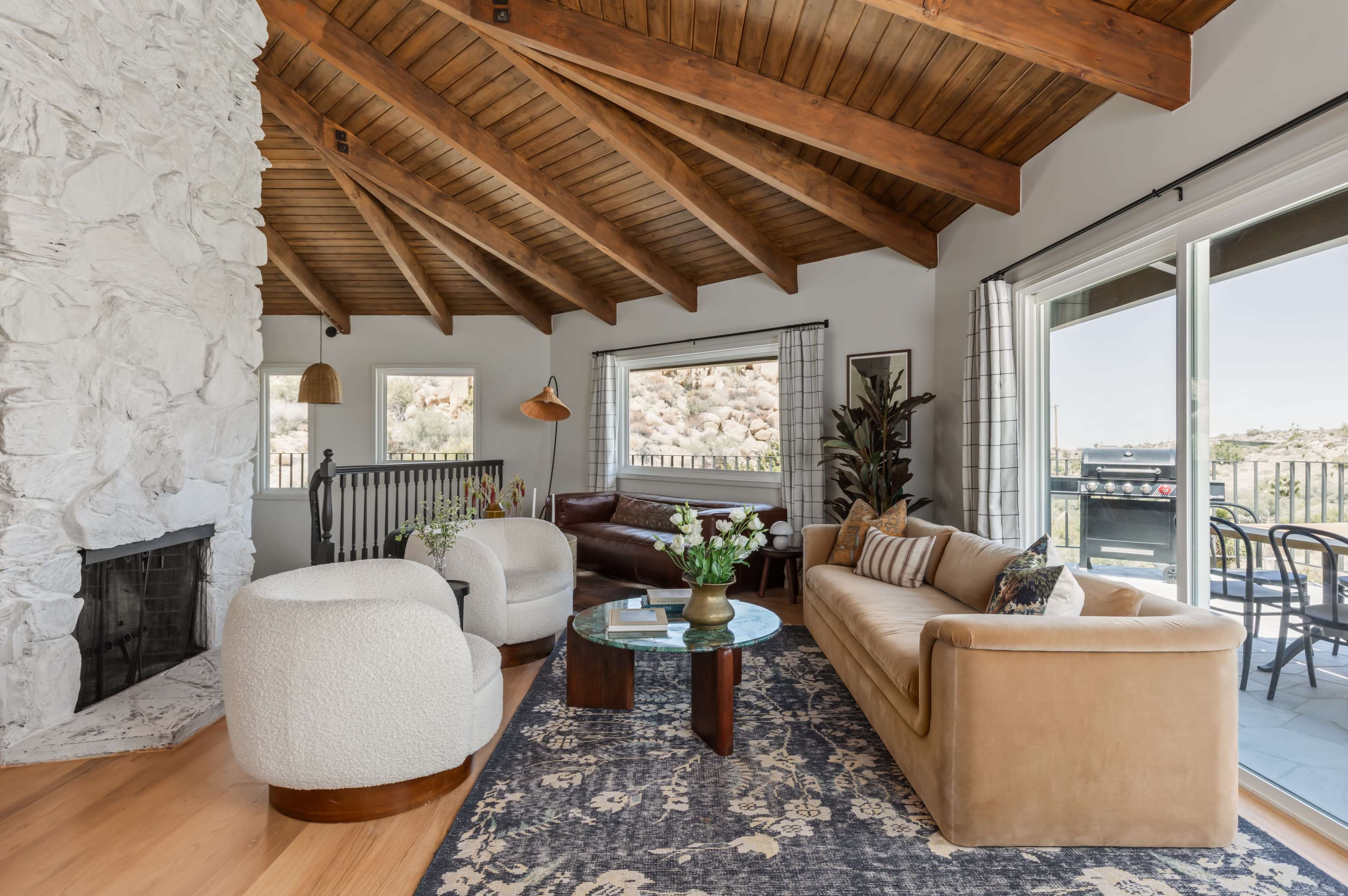 The image shows a cozy living room featuring a stone fireplace, wooden beams on the ceiling, and a mix of modern and traditional furniture.