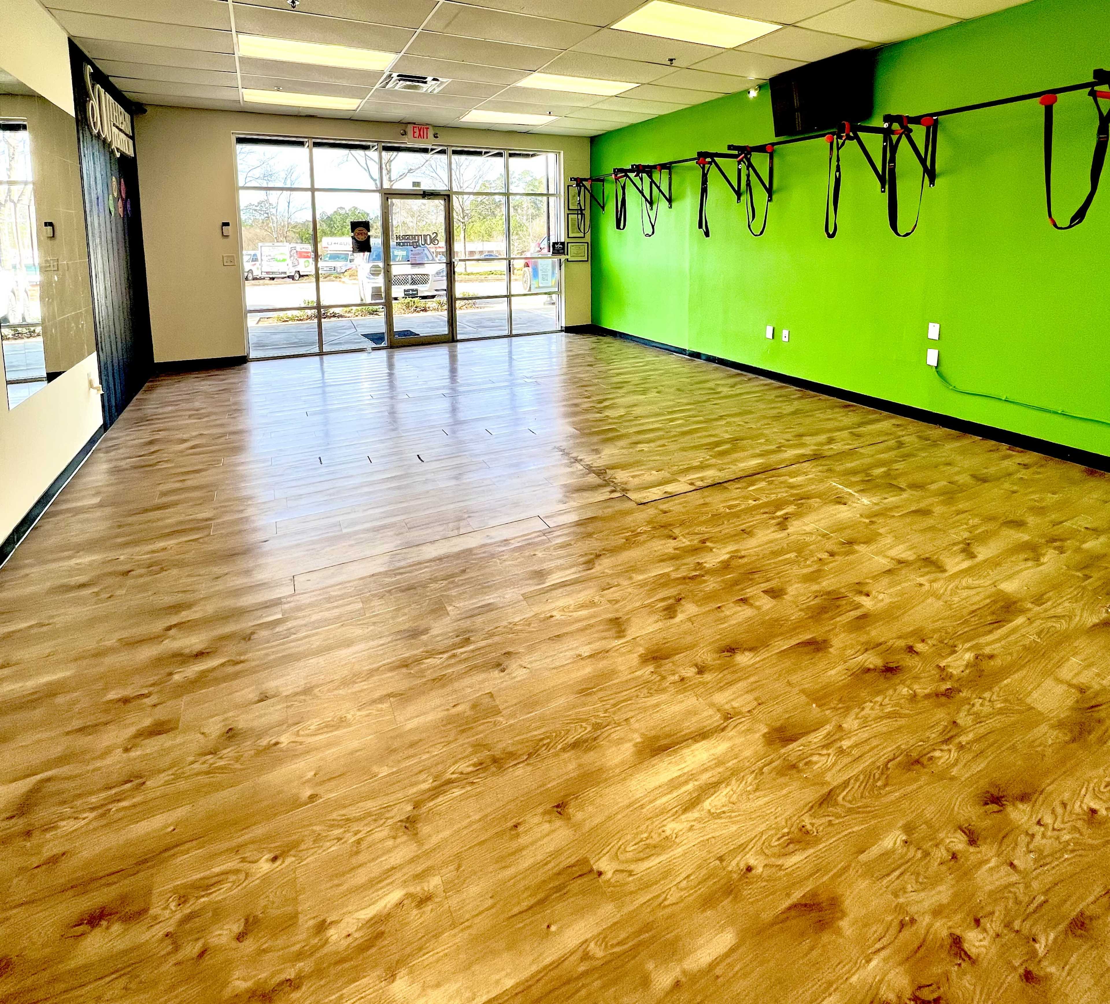 An empty fitness studio with wooden flooring, green walls, and suspension training equipment hanging on one side.