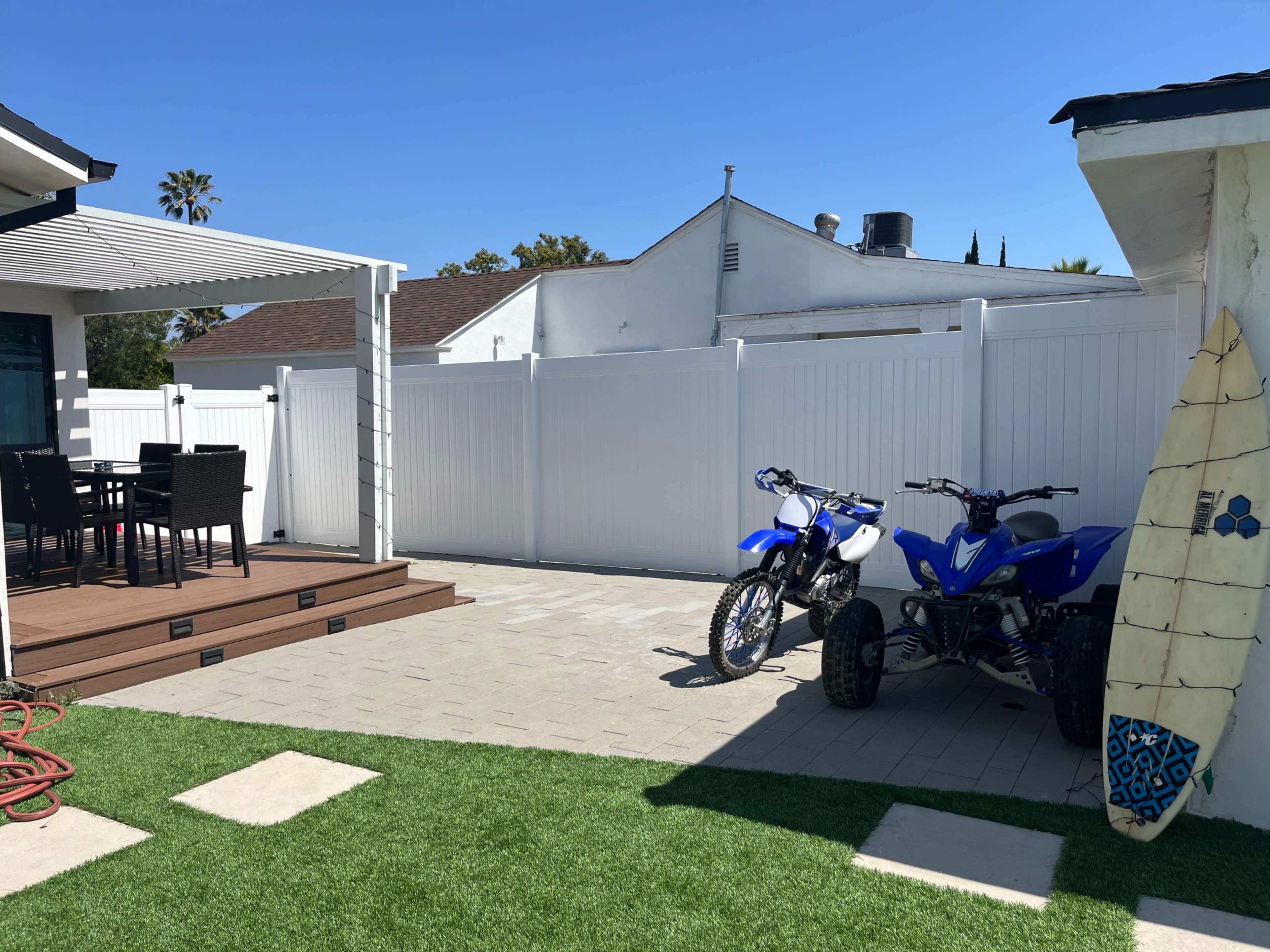 A backyard featuring a deck with black outdoor furniture, two blue ATVs parked on pavers, and a surfboard leaning against a white wall.